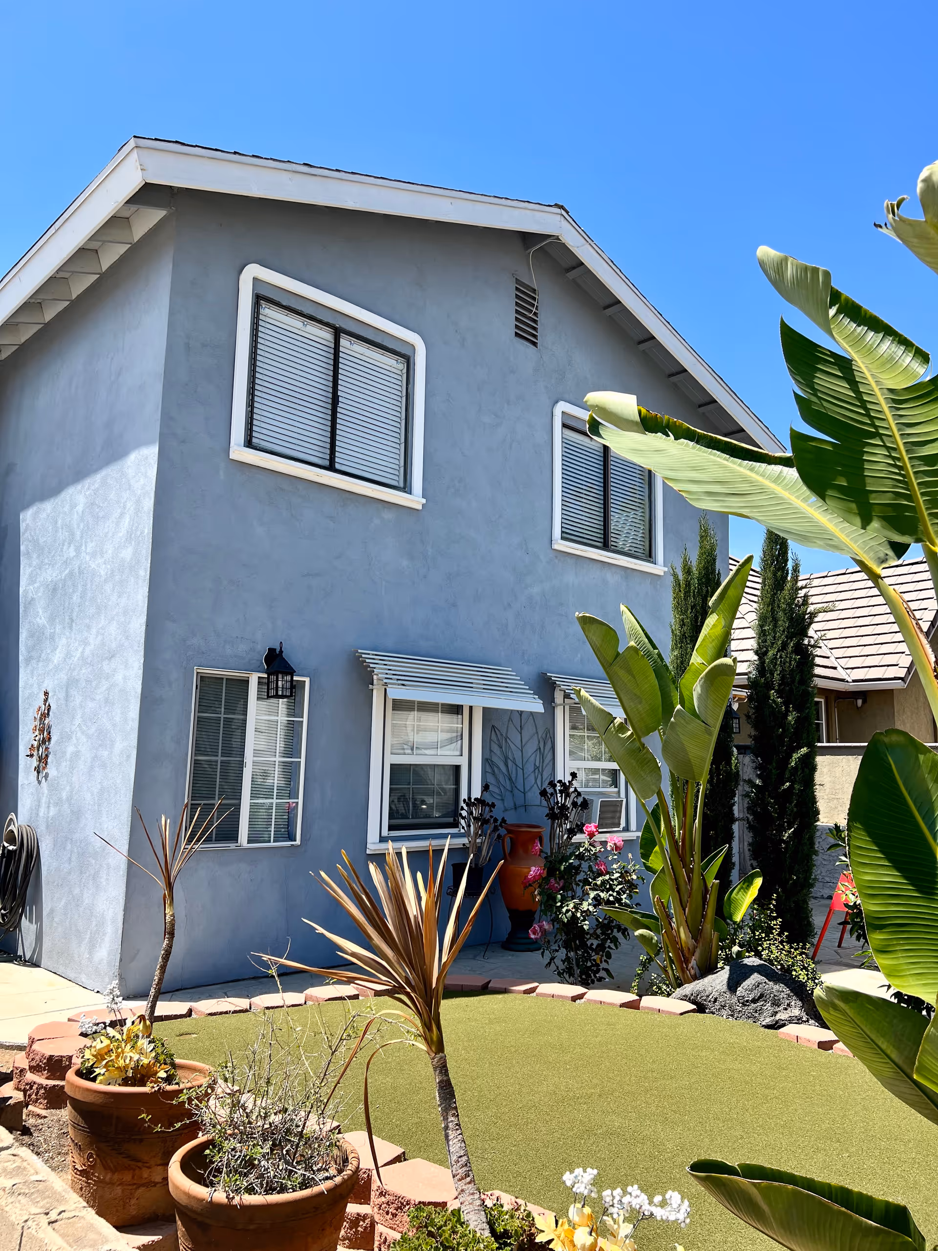 Front exterior of a two-story gray stucco house with windows, a small landscaped yard, potted plants, and large tropical leaves.