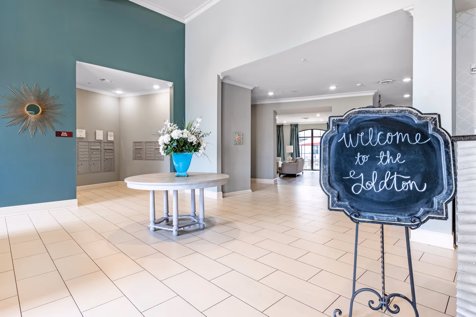 Interior view of a senior living facility lobby with a round table holding a blue vase filled with white flowers in the center. A decorative chalkboard sign on a stand reads 'welcome to the Goldton'. The walls are painted in teal and light gray, with mailboxes visible in a recessed area. The floor is tiled in a light beige color.