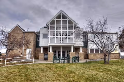 Exterior view of a two-story assisted living facility building with a large covered balcony, brick and siding walls, a patio area with green chairs and tables, leafless trees, and a grassy lawn under a cloudy sky.