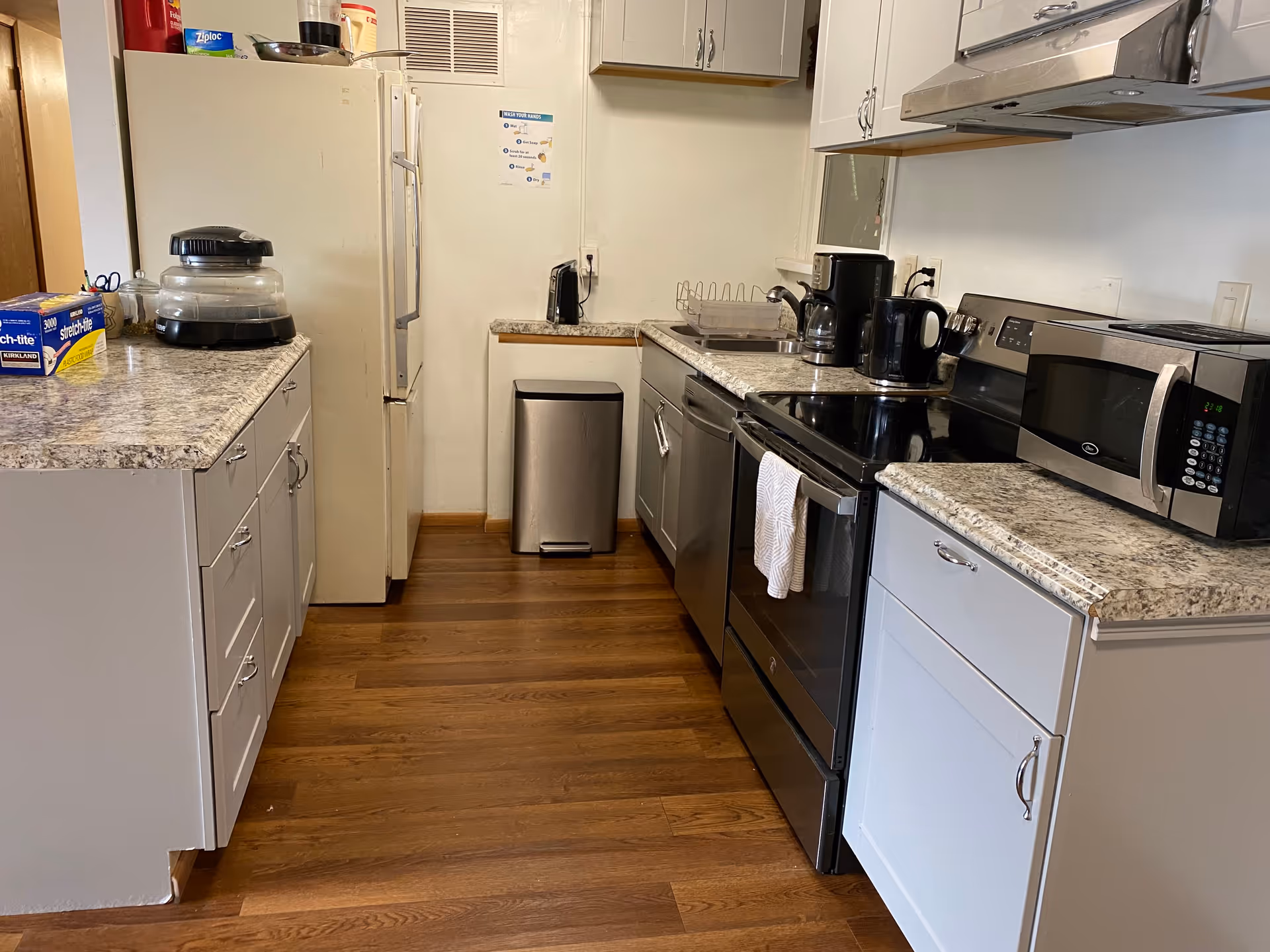 A clean kitchen with white cabinets and granite countertops. The kitchen features a refrigerator, a microwave, a stove with a towel hanging on the handle, a coffee maker, a kettle, a toaster, a dish drying rack, and a stainless steel trash can. The floor is wooden, and there is a small poster on the wall above the trash can.