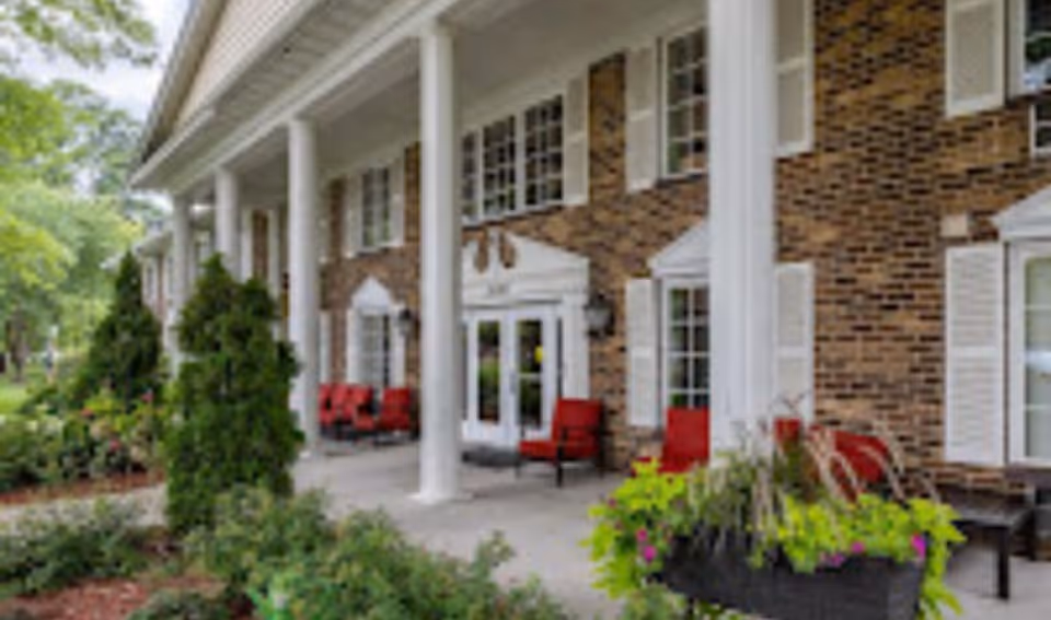 Exterior view of a brick building with white columns and multiple windows with white shutters. There is a covered porch area with red cushioned chairs and greenery including bushes and potted plants in the foreground.