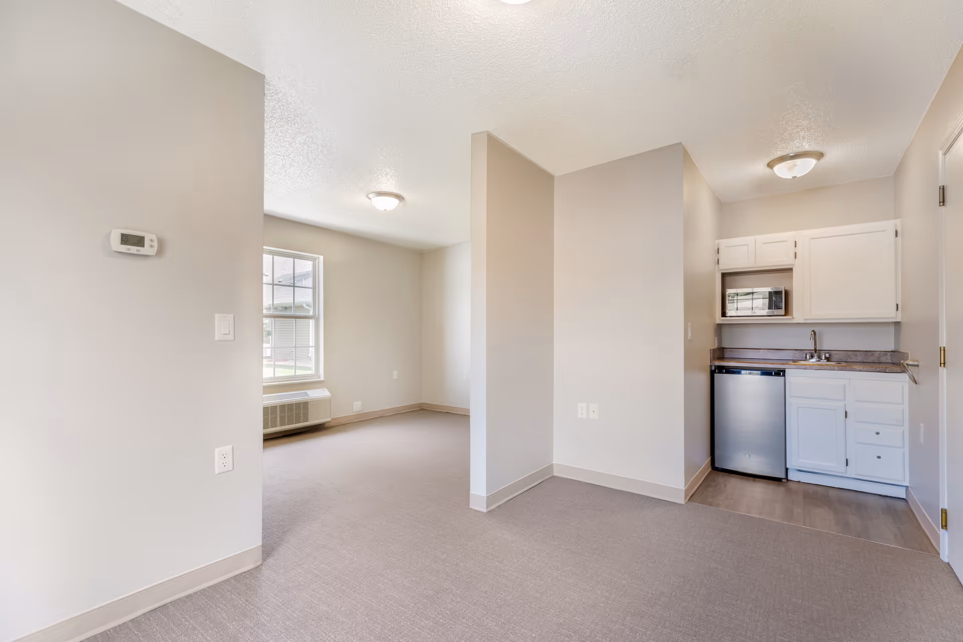 Interior view of a senior living facility apartment featuring a small kitchenette with white cabinets, a microwave, a mini refrigerator, and a sink. Adjacent to the kitchenette is an open area with beige carpet flooring, a window with natural light, and a wall-mounted thermostat.