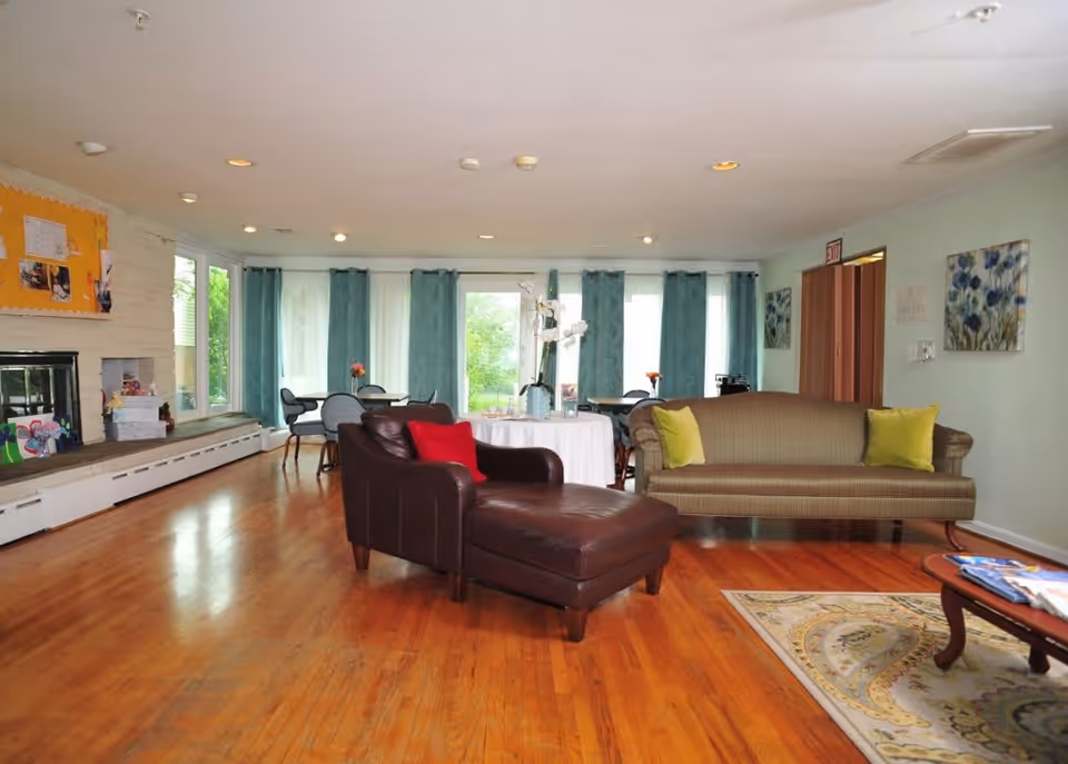 A spacious living room with wooden floors, a brown leather armchair with a red pillow, a striped sofa with two green pillows, a coffee table with magazines, and several round tables with chairs near large windows covered with teal curtains. The walls are light green with floral artwork, and there is a fireplace with a bulletin board above it on the left side.