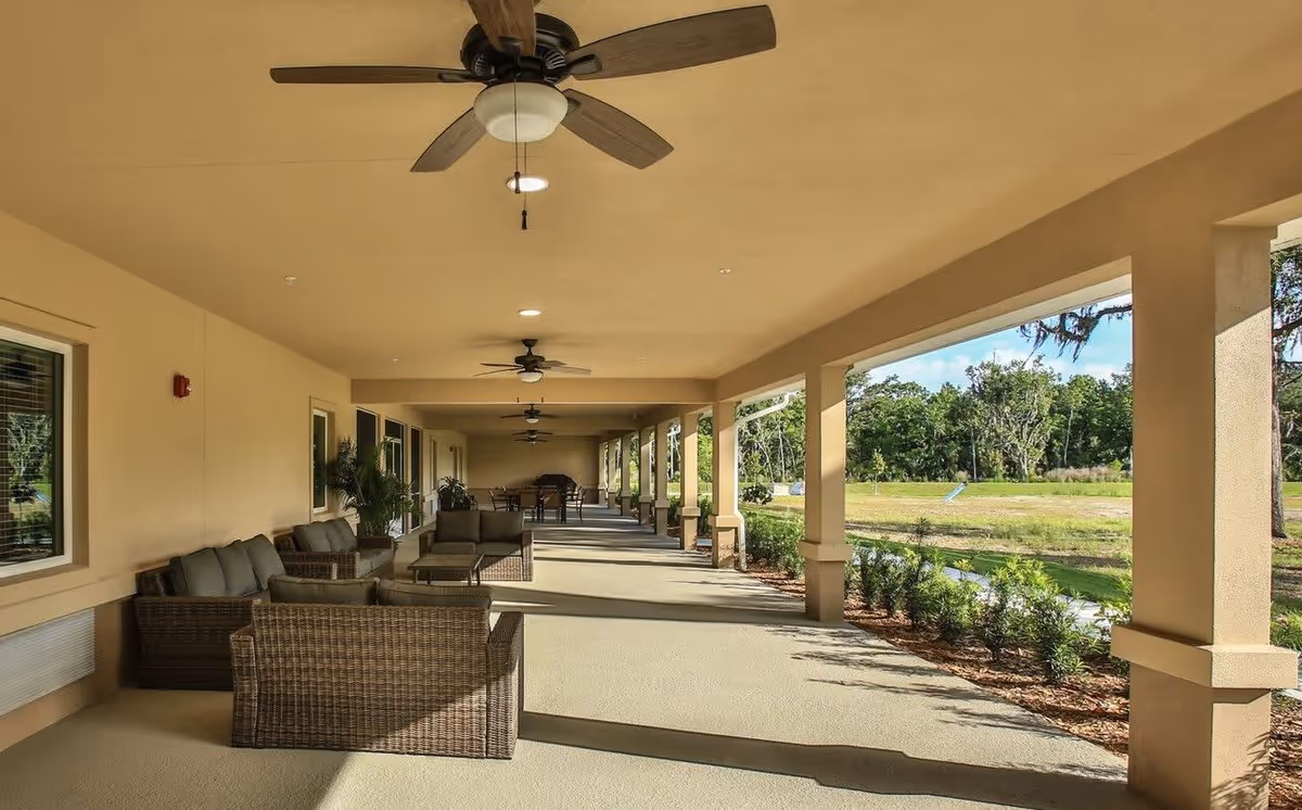 A spacious covered outdoor patio area at Ormond Manor with wicker seating arrangements including sofas and chairs with cushions. Ceiling fans are mounted on the ceiling, and the patio overlooks a green landscaped area with trees and shrubs.