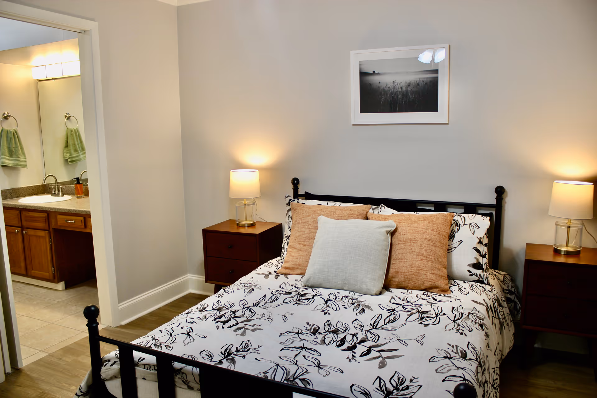 A furnished bedroom with a bed, two bedside tables and lamps, and an open doorway showing a bathroom sink.