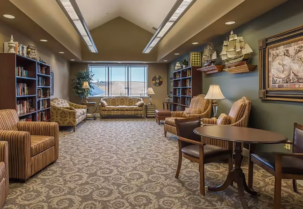 A spacious, well-lit living room with patterned carpet, multiple armchairs and sofas, two bookshelves filled with books, decorative model ships on wall shelves, table lamps, a round table with chairs, and a large window showing an outdoor view.