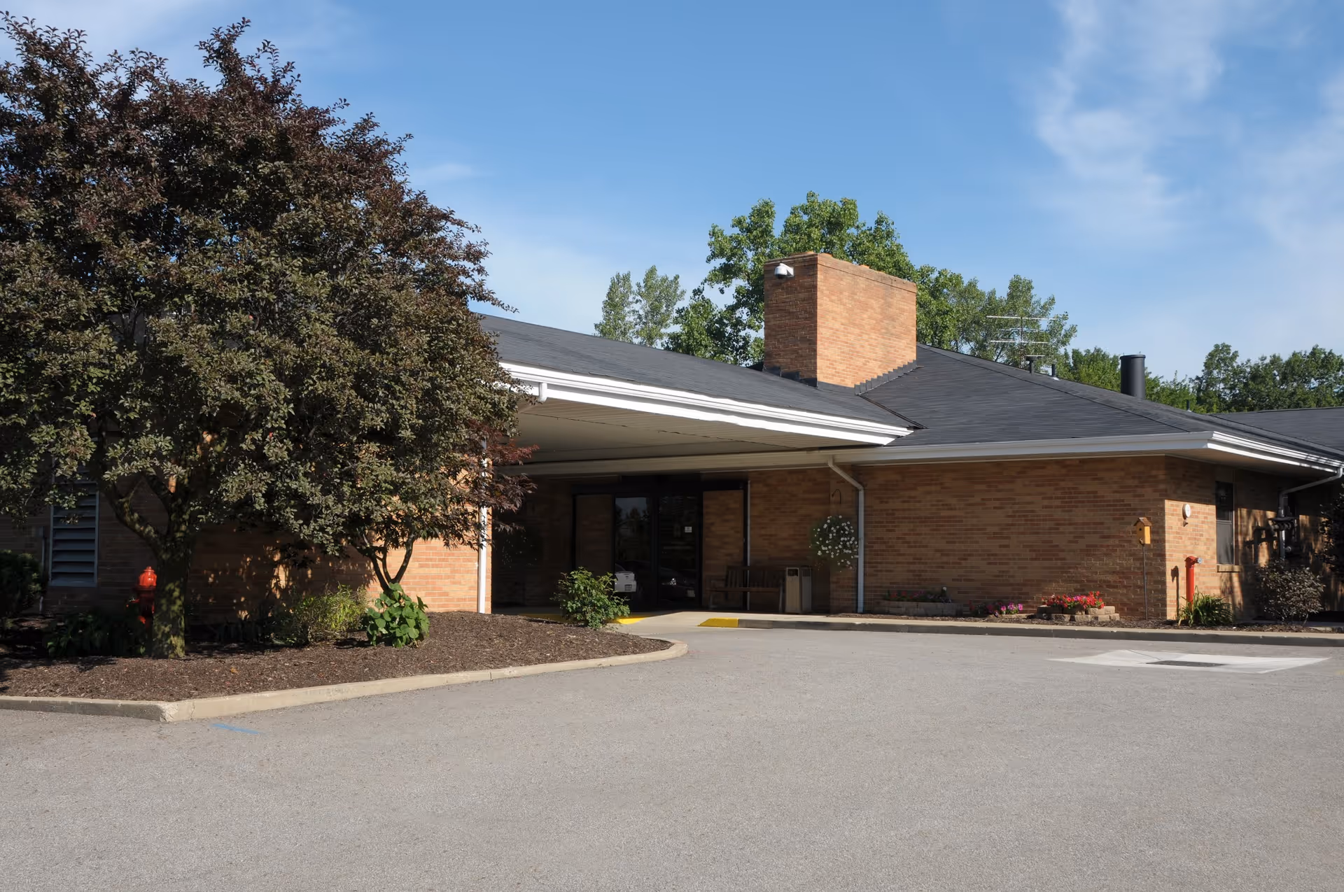 Front entrance of a one-story brick building with a covered porte-cochère, trees, and a driveway.