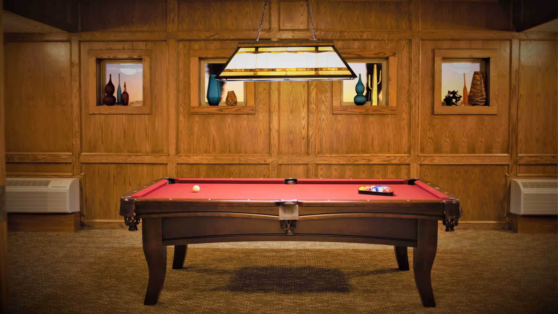 A pool table with a red felt surface in a room with wood-paneled walls. The pool table has a rack of billiard balls and a cue ball on it. Above the table hangs a rectangular light fixture. The walls have built-in display niches with decorative vases and sculptures. Two air conditioning units are visible on either side of the room.