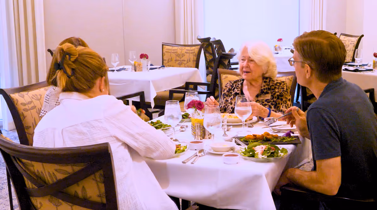Four people sitting around a dining table in a well-lit room, engaged in conversation while having a meal. The table is set with plates of food, glasses of water, and silverware. The chairs have patterned upholstery and the room has white walls and curtains.