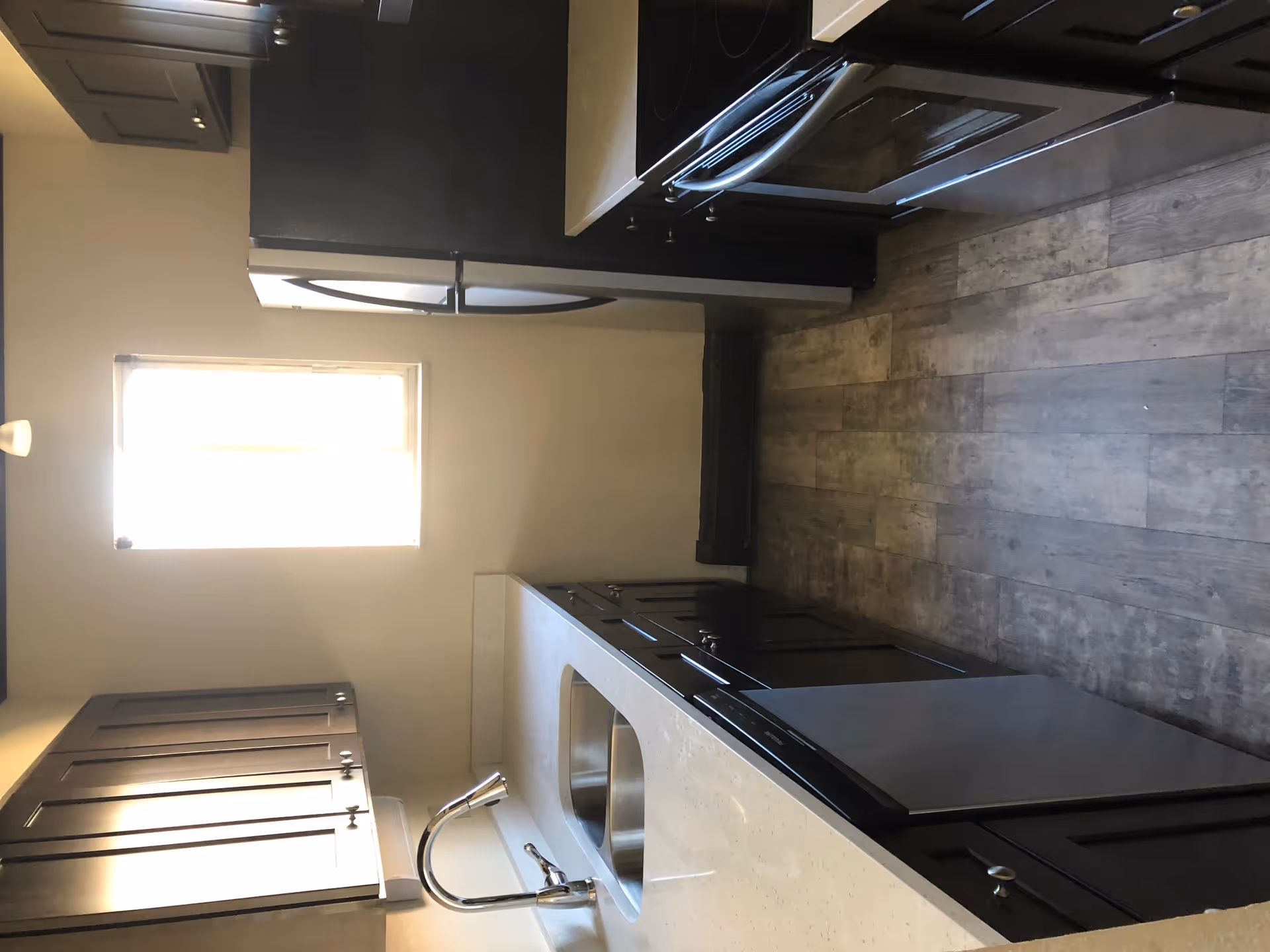 A narrow kitchen with dark wooden cabinets, a stainless steel double sink, dishwasher, refrigerator, and electric stove. There is a small window at the end of the kitchen letting in natural light, and the floor has a wood-like finish.