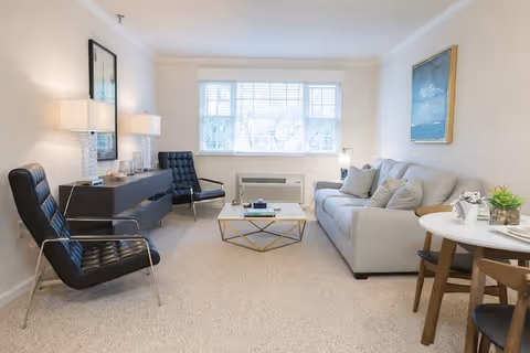 A bright and cozy living room with a large window letting in natural light. The room features a light gray sofa with several cushions, two black modern armchairs, a glass coffee table with a gold geometric frame, a dark wooden sideboard with two table lamps, and a small round dining table with two chairs. A framed painting hangs on the wall above the sofa.