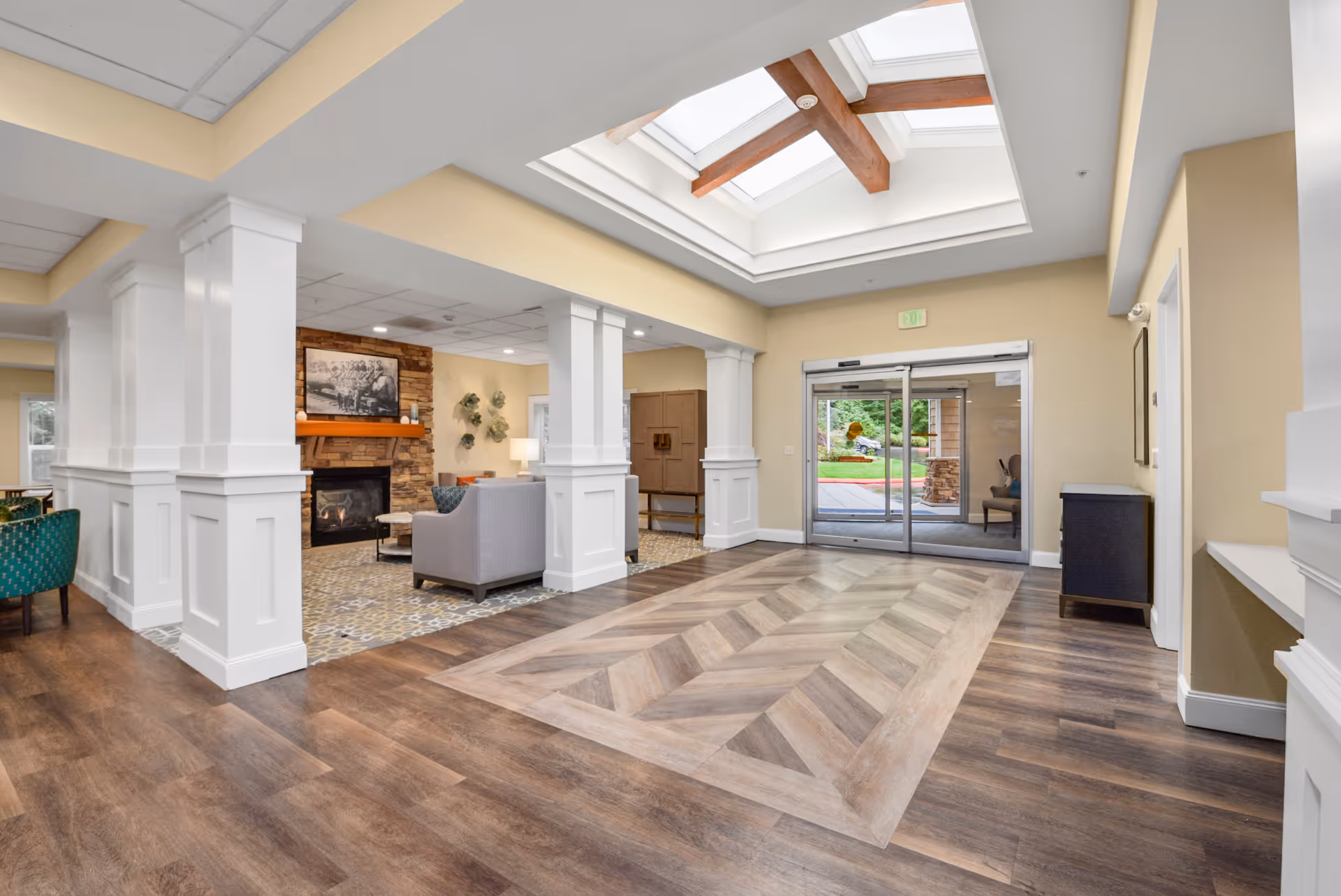 Interior view of a senior living facility lobby area with a skylight ceiling, wood and tile flooring, white pillars, a seating area with chairs and a fireplace, and glass automatic doors leading outside.
