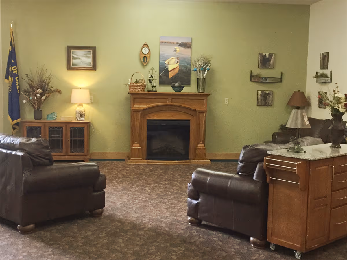 A cozy living room area with two brown leather armchairs facing a wooden fireplace. The walls are painted light green and decorated with various paintings and a clock. There is a wooden cabinet with a lamp and decorative items on the left side, and a wooden rolling cart with a granite top and a lamp on the right side. The floor is carpeted with a patterned design.