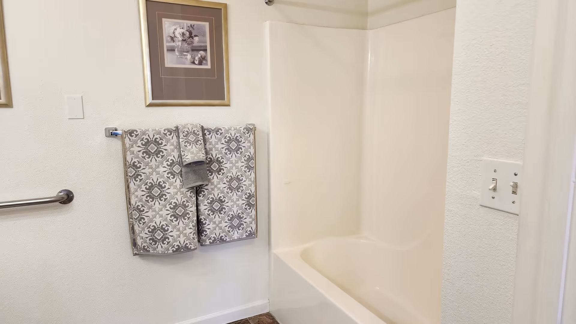 A bathroom with a white bathtub and shower combination. Two patterned towels and a smaller hand towel hang on a towel rack on the wall. Above the towels is a framed floral picture. There is a metal grab bar on the left wall and a light switch panel on the right wall.