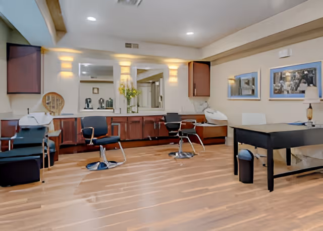 Interior view of a salon area in a senior living facility with wooden flooring, salon chairs, mirrors on the wall, and a desk with a lamp and chair. The room is well-lit with wall sconces and has framed artwork on the walls.
