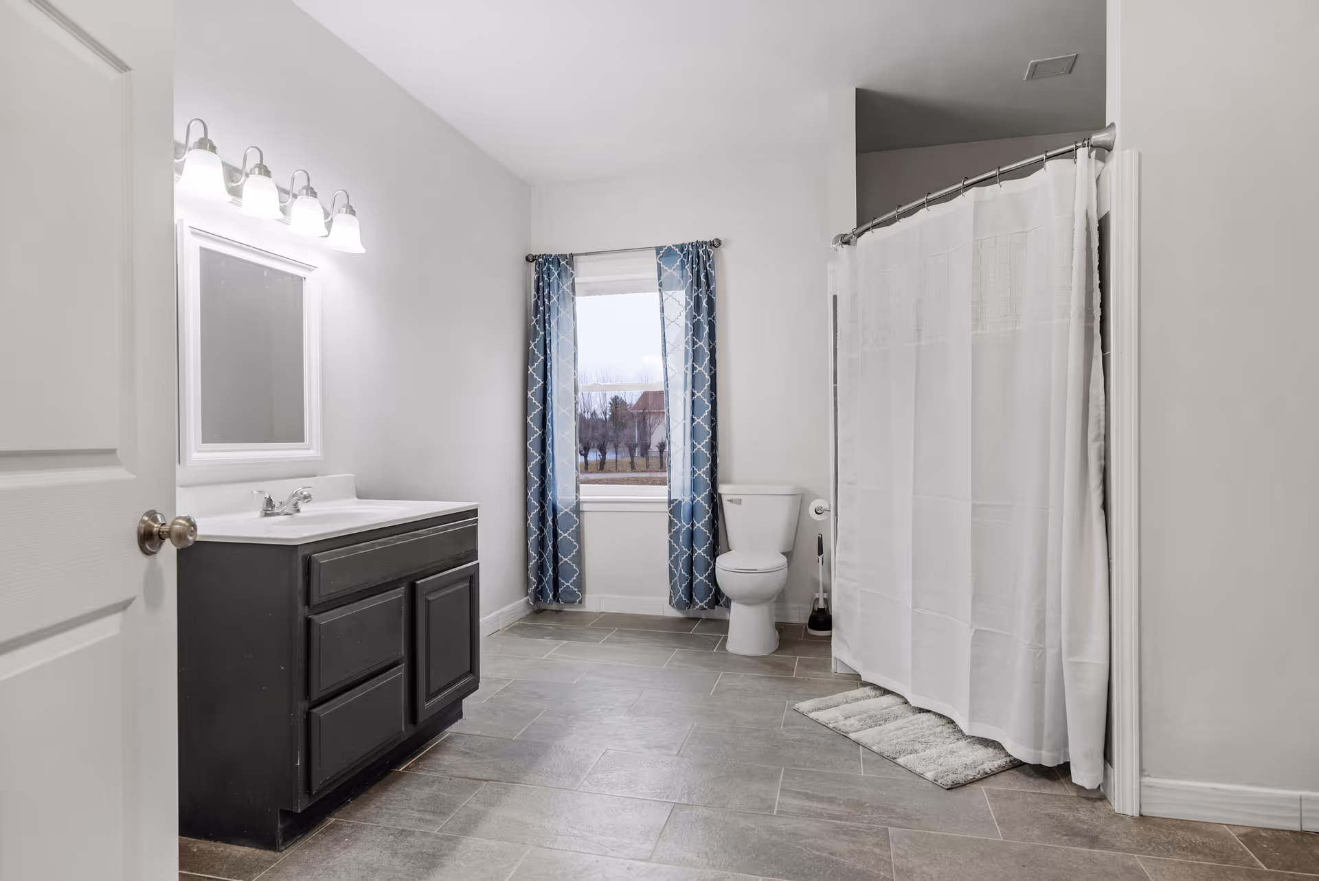 A spacious bathroom with a black vanity cabinet and white countertop, a large mirror with four light fixtures above it, a toilet next to a window with blue patterned curtains, and a shower area with a white curtain and a small rug on the floor.