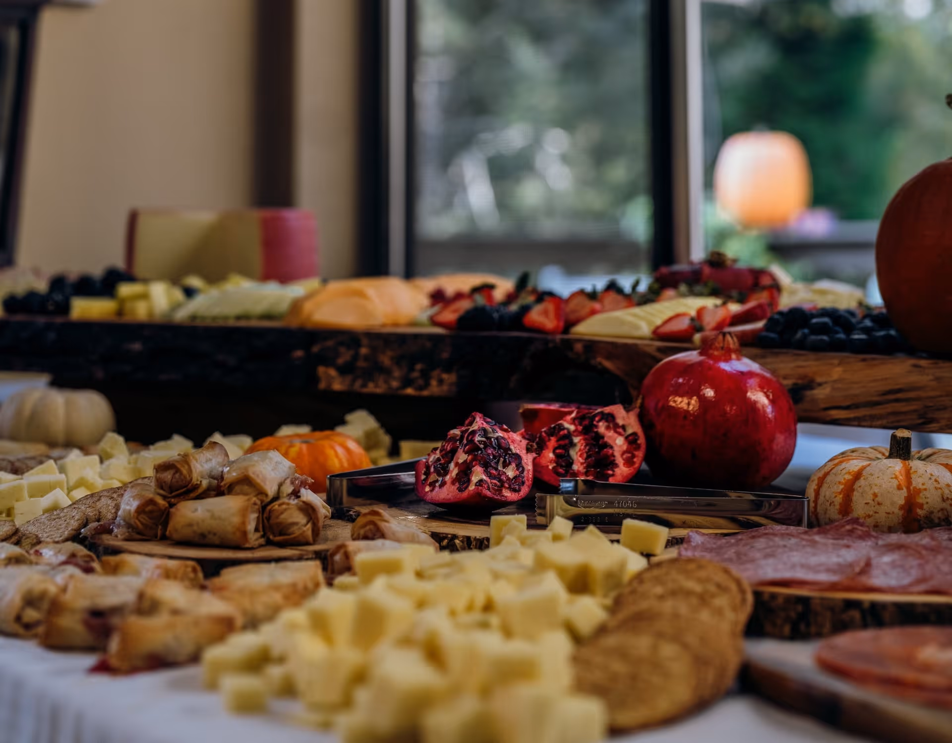 A close-up of a buffet table indoors with cheeses, crackers, pomegranate, fruit and various appetizers arranged on platters.