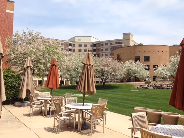 Outdoor patio area with several tables and chairs, each table shaded by closed umbrellas in beige and rust colors. The patio overlooks a well-maintained grassy area with blooming trees and a multi-story building in the background under a partly cloudy sky.