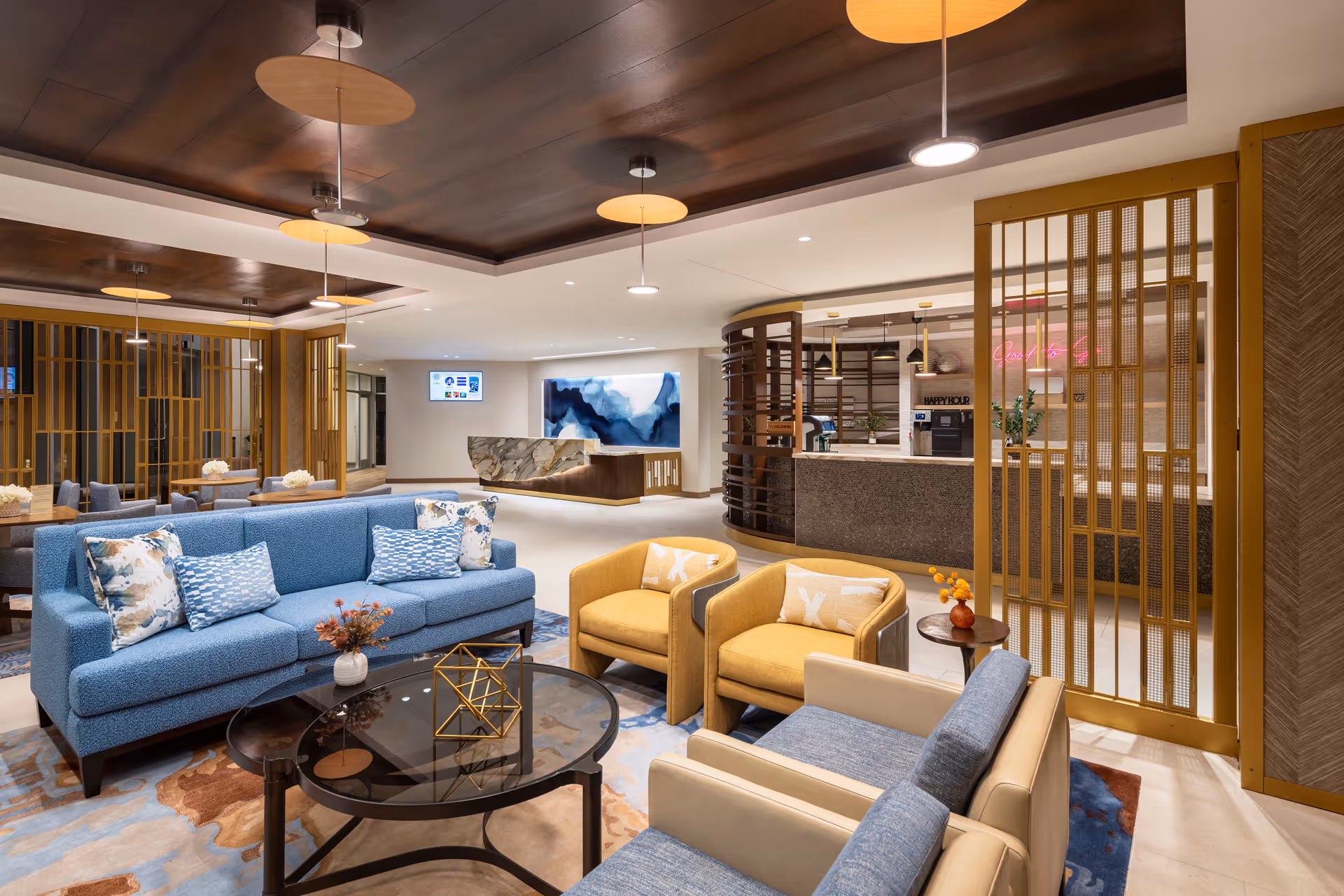 A modern senior living facility lounge area with blue and beige sofas, yellow armchairs, a glass coffee table with decorative items, and a reception desk in the background. The space features warm lighting, wooden ceiling panels, and gold-colored room dividers.