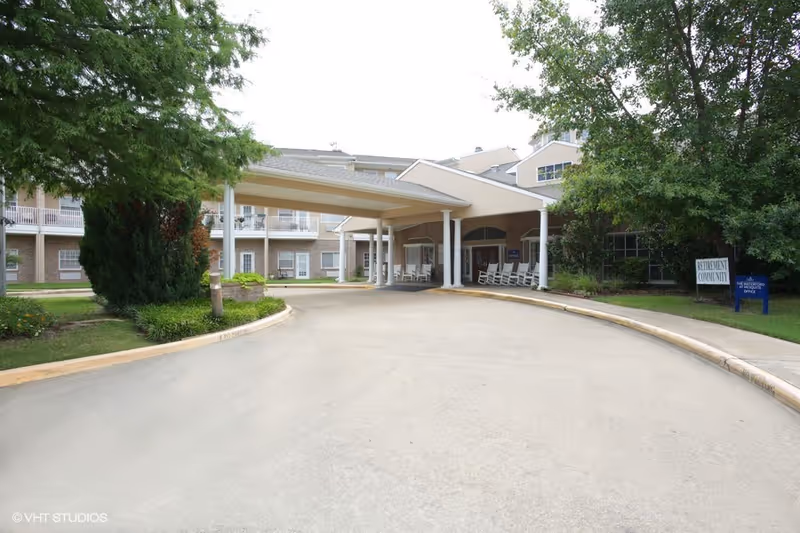 Entrance driveway and covered drop-off area of a senior living facility with rocking chairs on the porch, surrounded by trees and greenery. Signs indicate it is a retirement community and the Mesquite office.