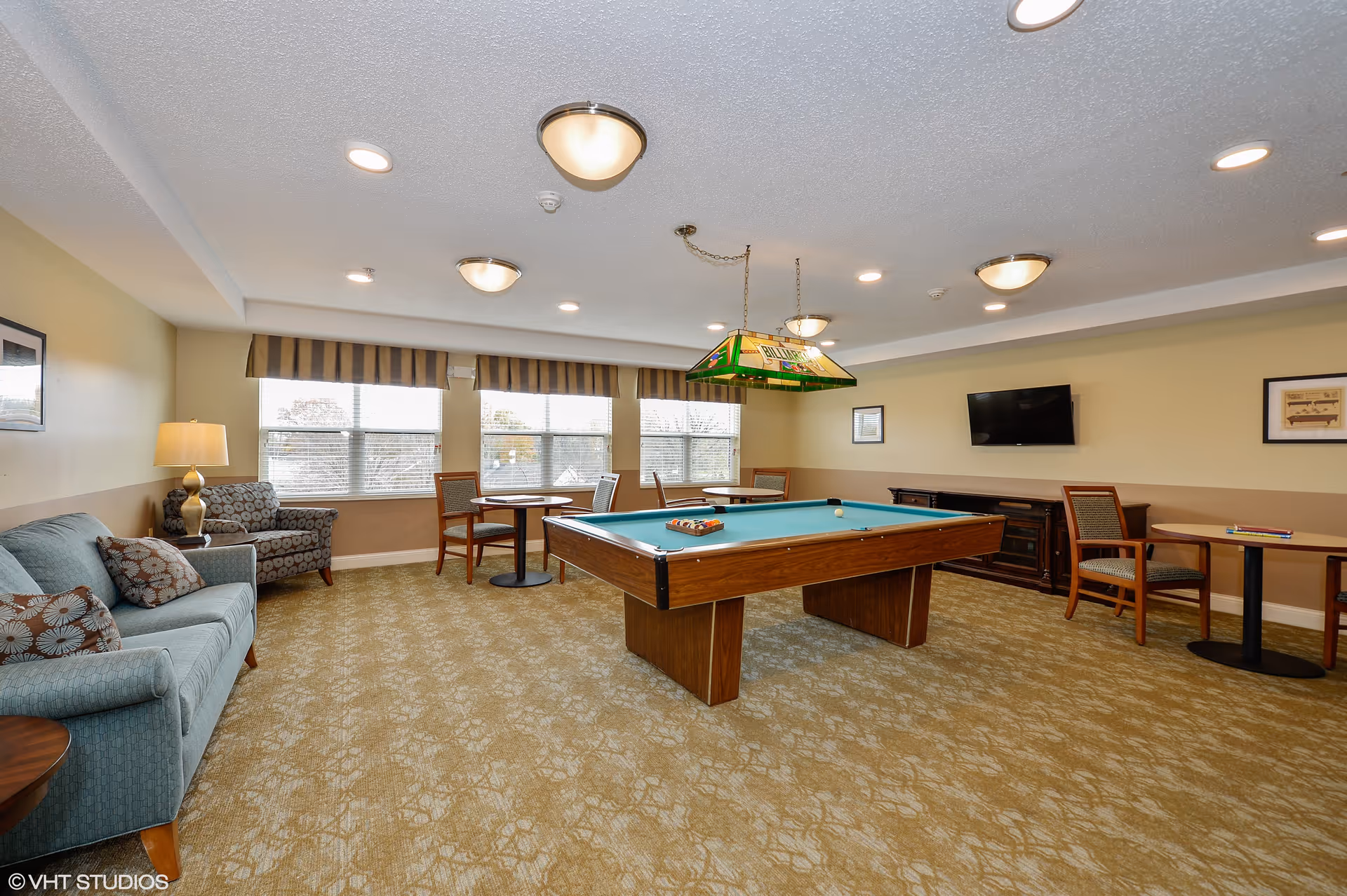A bright common room with a pool table in the center, seating areas, small tables, and a wall-mounted TV.
