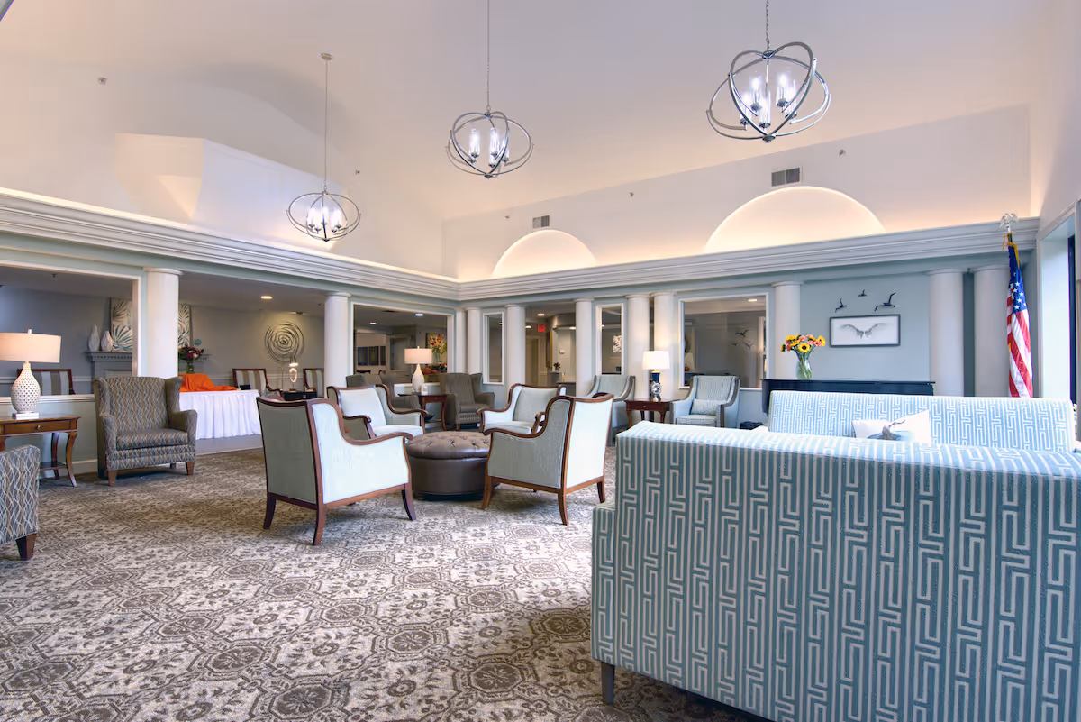 Spacious senior living community lounge area with patterned carpet, multiple armchairs and sofas arranged around a central ottoman, decorative lamps on side tables, and modern chandeliers hanging from a high ceiling. The room features white columns, soft lighting, artwork on the walls, and an American flag in the corner.