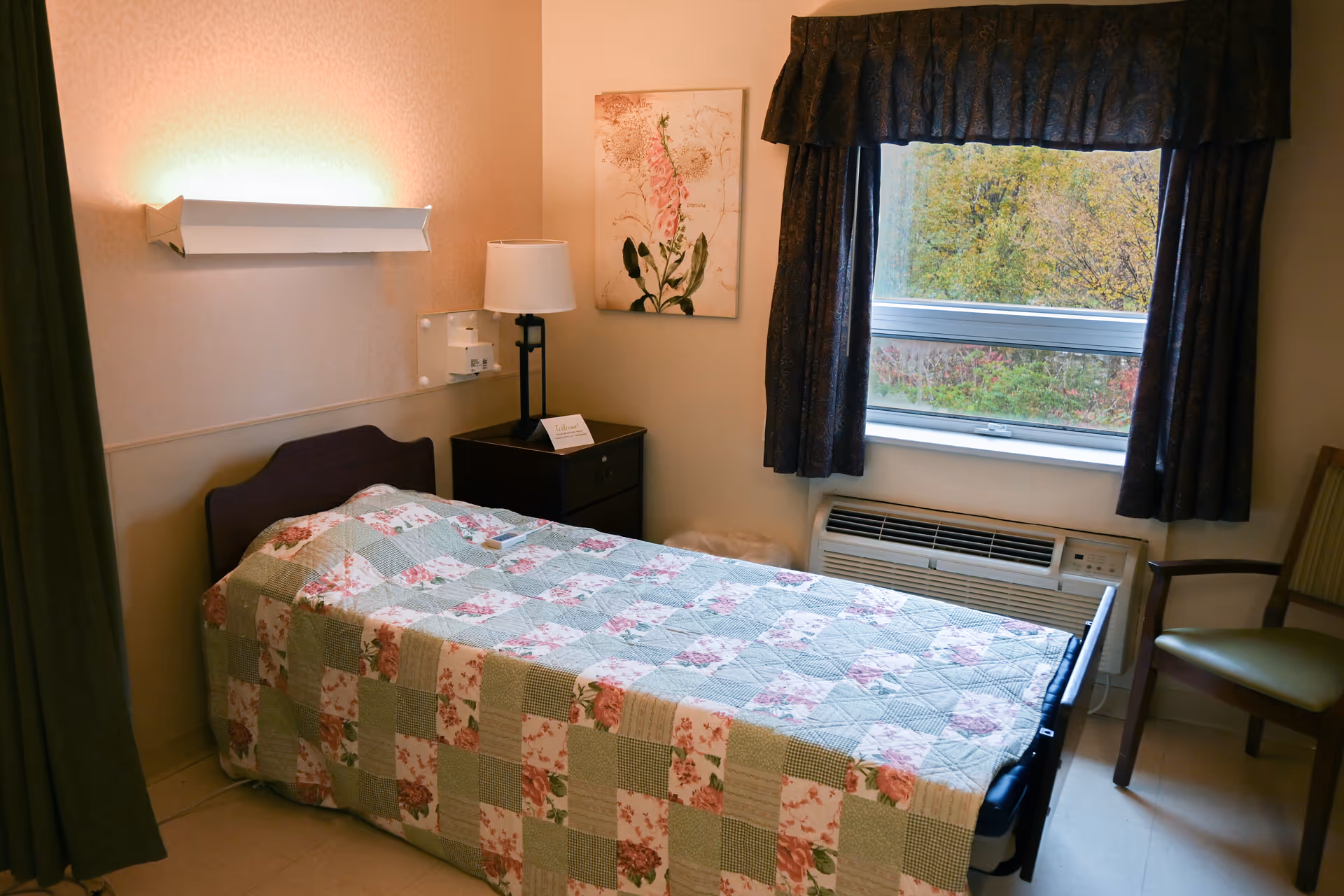 A small bedroom in a senior living facility with a single bed covered by a patchwork quilt in green, pink, and white floral patterns. Next to the bed is a dark wooden nightstand with a lamp and a small card. On the wall above the nightstand is a floral painting. A window with dark curtains shows trees outside, and below the window is an air conditioning unit. A wooden chair with a green cushion is placed near the window.