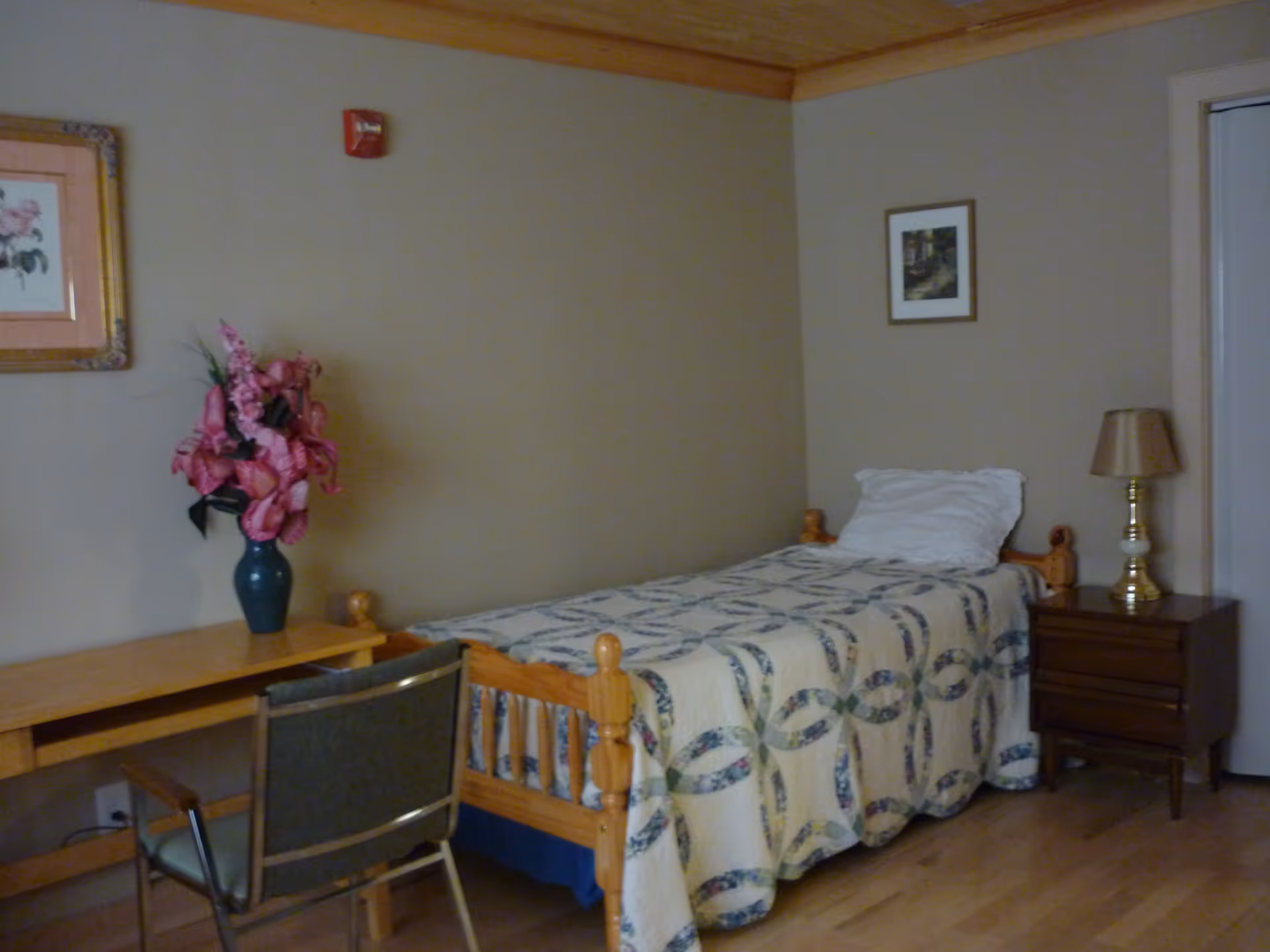 A simple bedroom with a single wooden bed covered with a patterned quilt and a white pillow. Next to the bed is a wooden nightstand with a brass lamp. On the left side, there is a wooden desk with a vase of pink flowers and a chair. The walls are beige with two framed pictures hanging, and the floor is wooden.