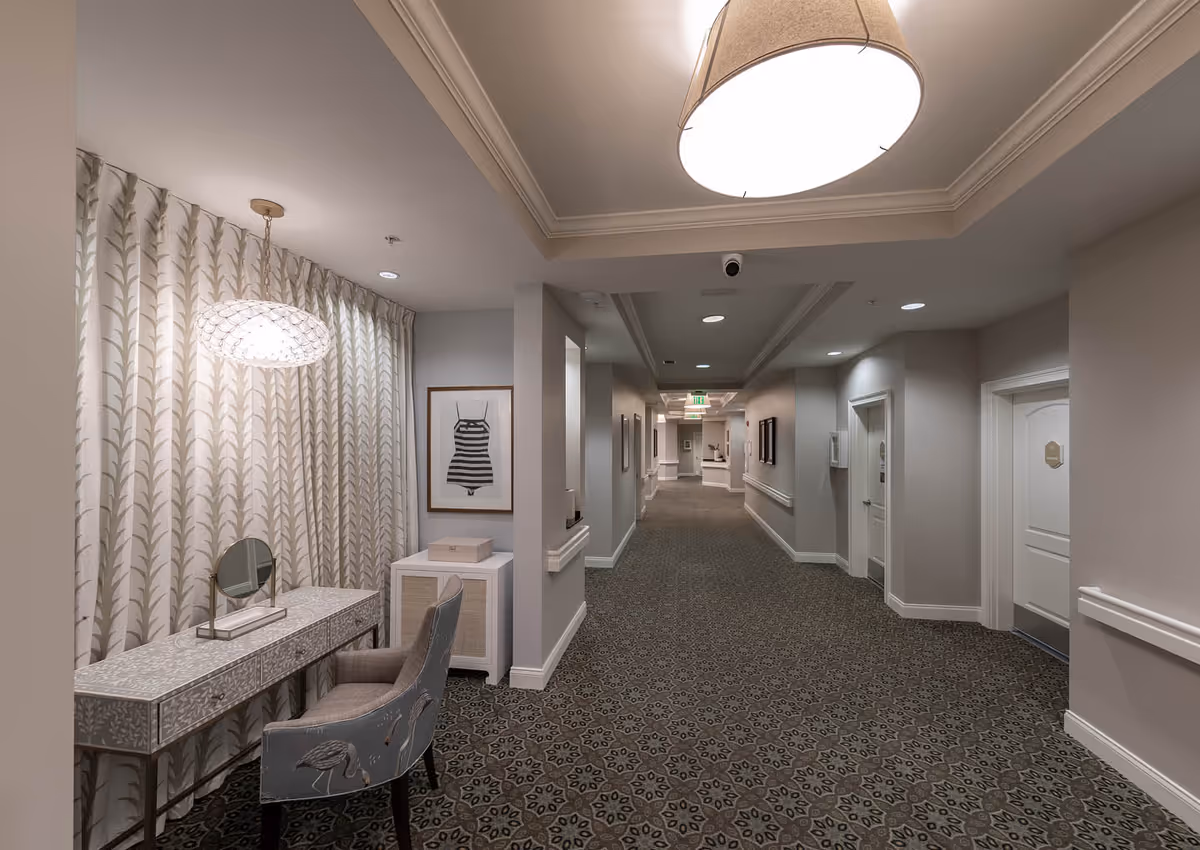 A well-lit hallway in a senior living facility with patterned carpet and light gray walls. On the left side, there is a decorative vanity table with a mirror and a cushioned chair featuring a flamingo design. A framed picture of a striped swimsuit hangs on the wall above a small cabinet. The hallway extends into the distance with several doors and ceiling lights.