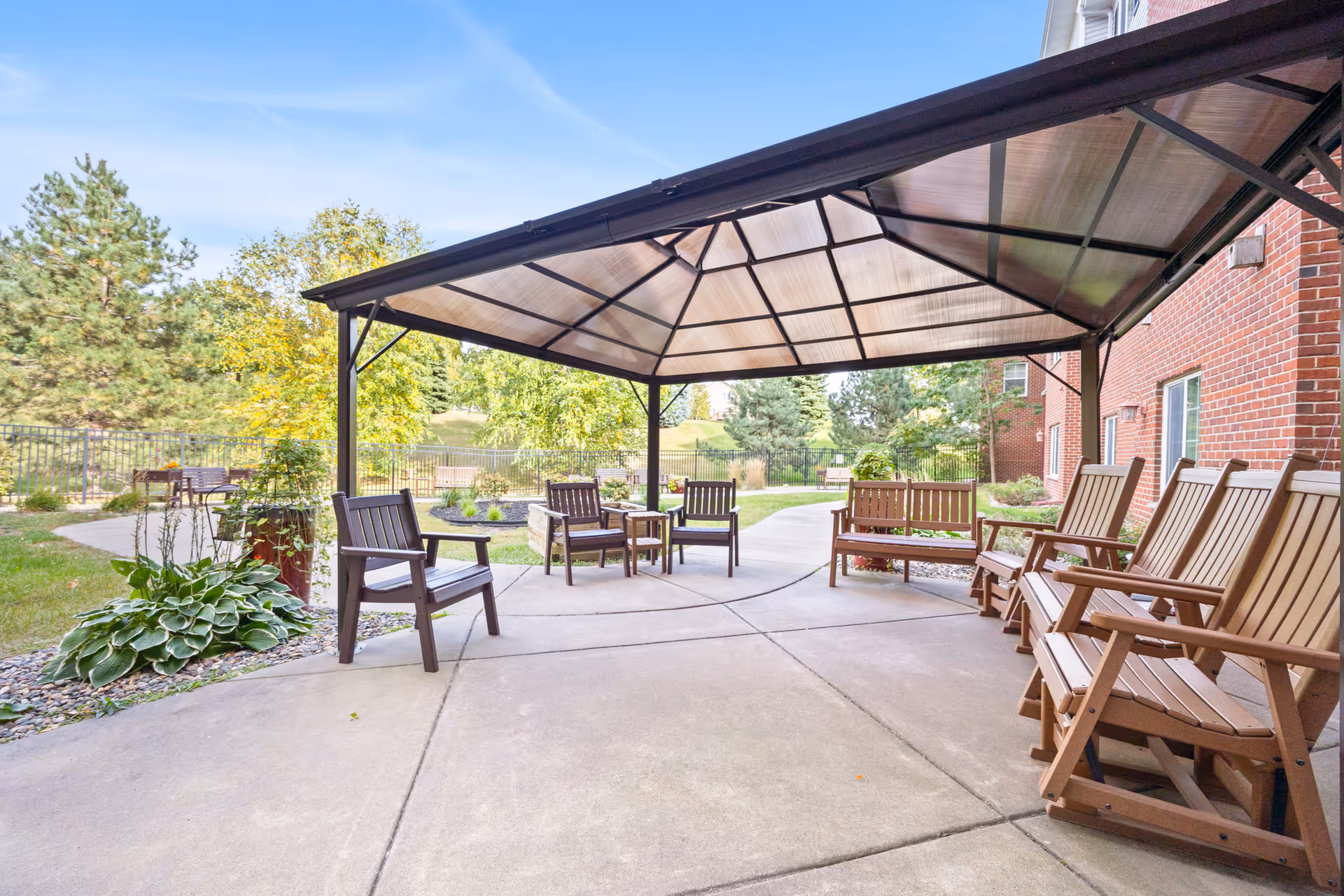 Outdoor covered patio area with several wooden chairs and benches arranged around a concrete floor. The patio is adjacent to a red brick building and surrounded by greenery including trees, shrubs, and a lawn. The sky is clear and blue.