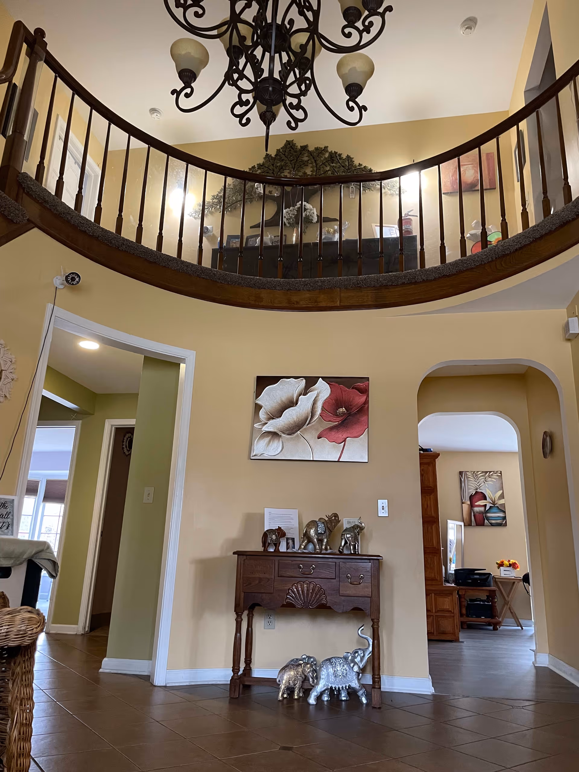 Interior view of a senior living facility showing a two-story area with a curved wooden railing on the upper level. Below, there is a small wooden table with decorative elephant figurines on and underneath it. Above the table hangs a painting of two large flowers, one white and one red. The walls are painted beige, and there are two doorways leading to other rooms, one with green walls and the other with beige walls and additional artwork visible. A chandelier with multiple lights hangs from the ceiling.