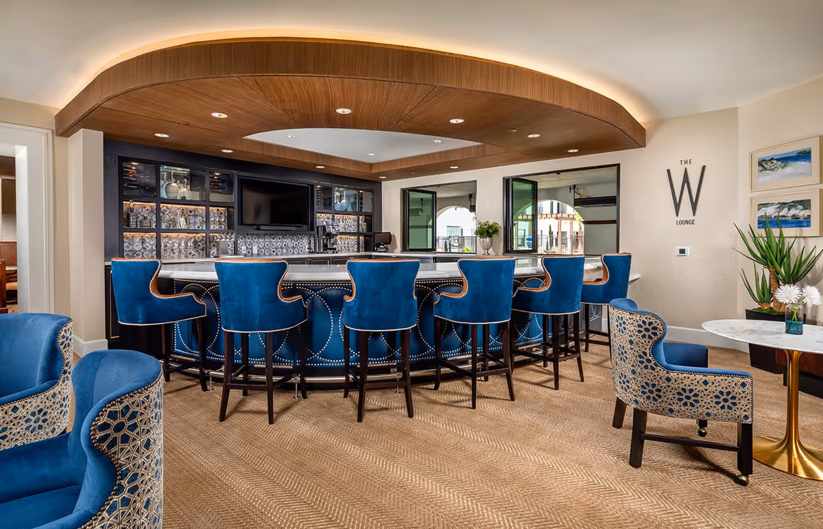 A stylish lounge area featuring a curved bar with blue upholstered bar stools and a wooden ceiling accent. The bar has a marble countertop and a backdrop with shelves and a television. There are additional blue and patterned chairs around small tables, with plants and framed artwork on the walls. The wall displays the text 'The W Lounge'.