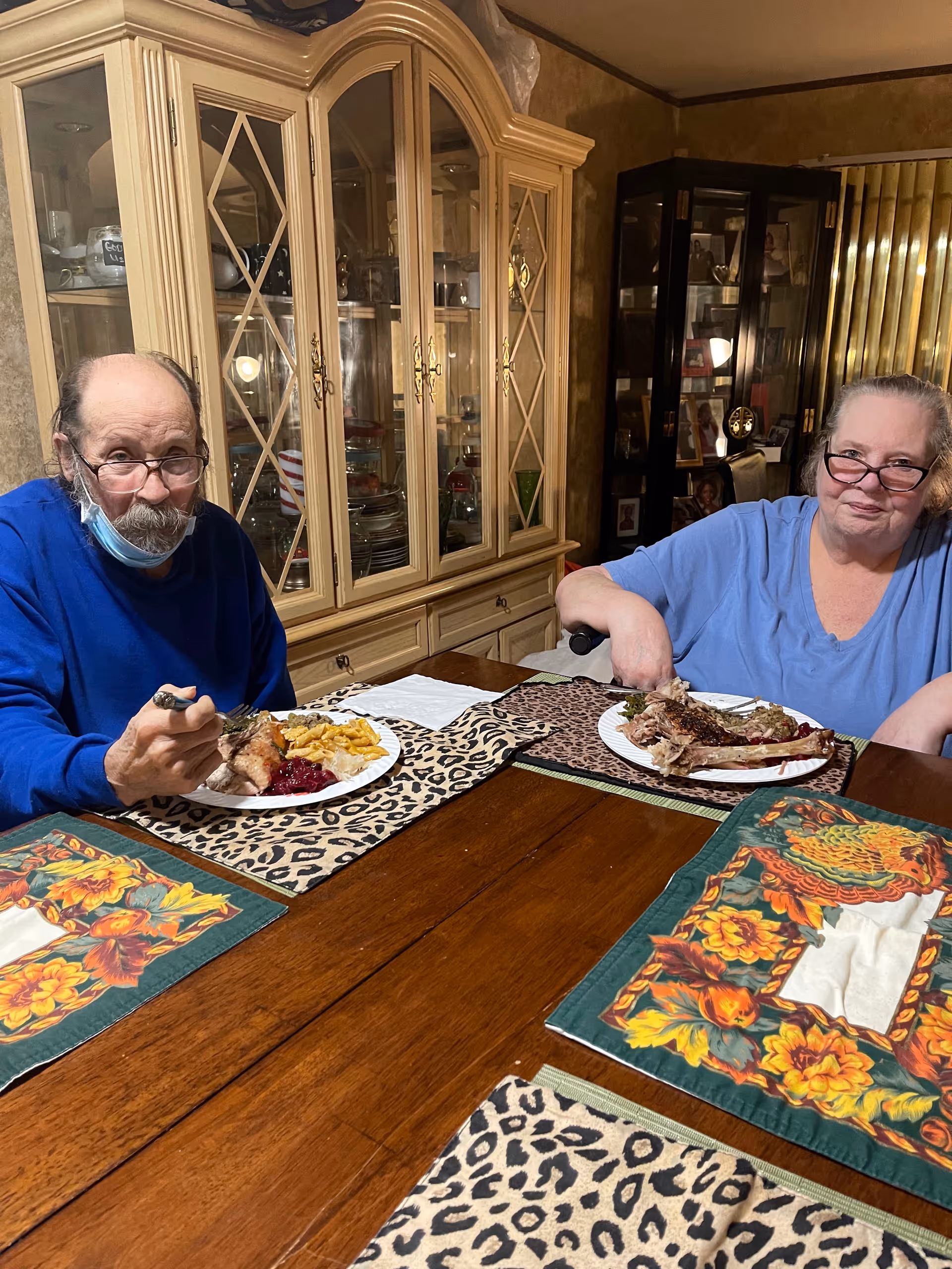 Two elderly people sitting at a wooden dining table with colorful placemats, eating a meal that includes turkey, macaroni and cheese, and cranberry sauce. Behind them is a large glass-fronted cabinet filled with dishes and glassware, and another cabinet with framed photos and decorative items.