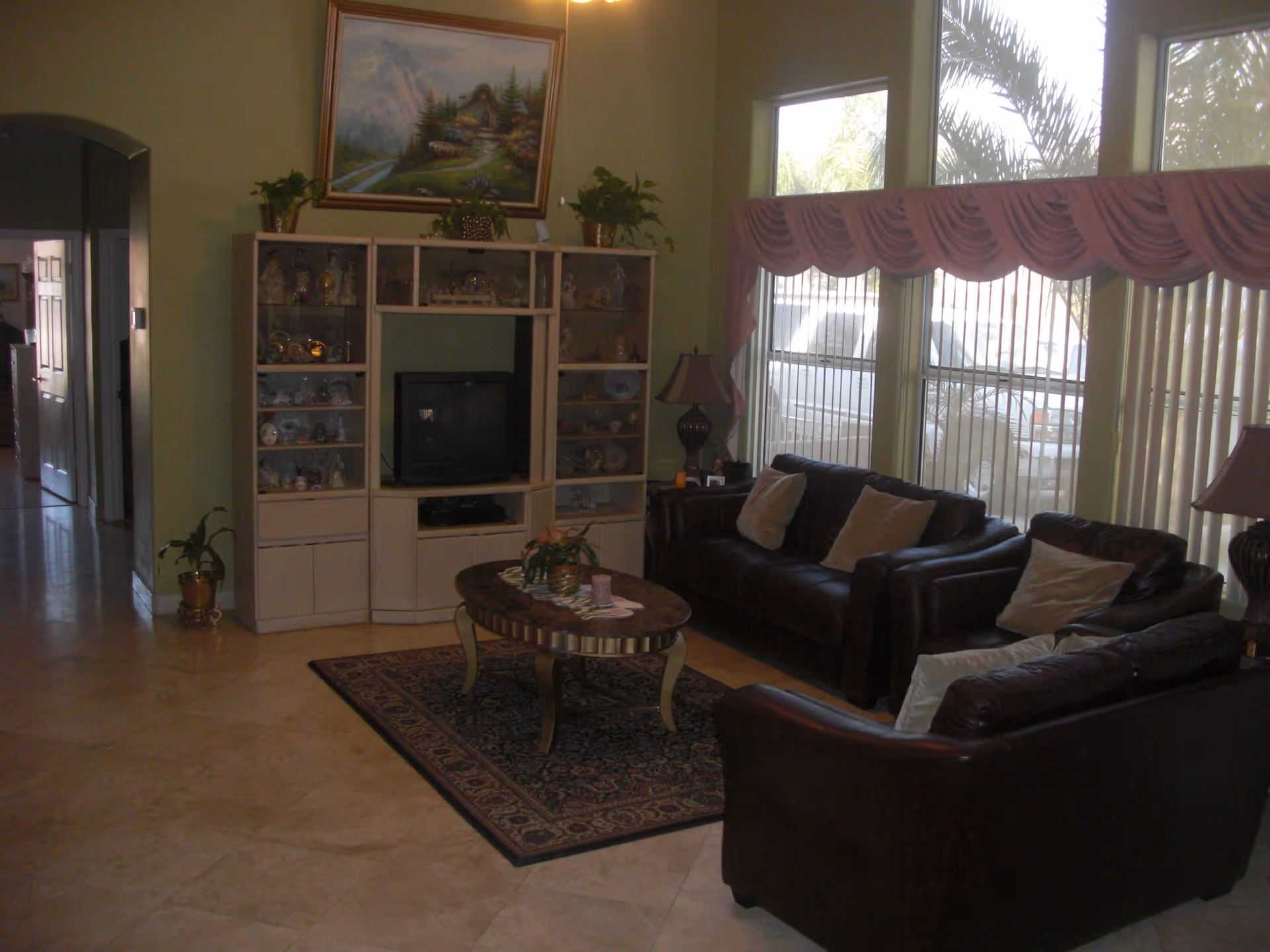 A living room with two dark brown leather sofas arranged around a wooden coffee table on a patterned rug. There is a white entertainment center with a TV and decorative items, and a large painting above it. The room has large windows with vertical blinds and pink valances, and two table lamps on either side of the sofas. The floor is tiled and there are some potted plants in the room.