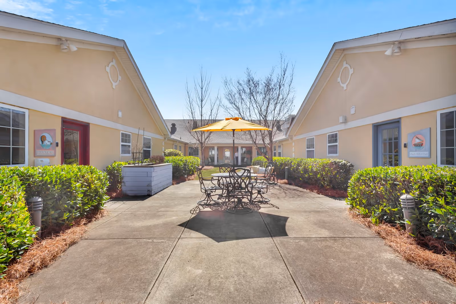 Outdoor courtyard area between two beige buildings with a concrete walkway, green bushes along the sides, a round metal table with four chairs, and a yellow umbrella providing shade. The sky is clear and blue.