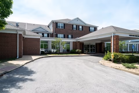 Front exterior view of a senior living facility building with a covered entrance, brick walls, multiple windows, and a driveway leading up to the entrance. There are some shrubs and greenery around the building.