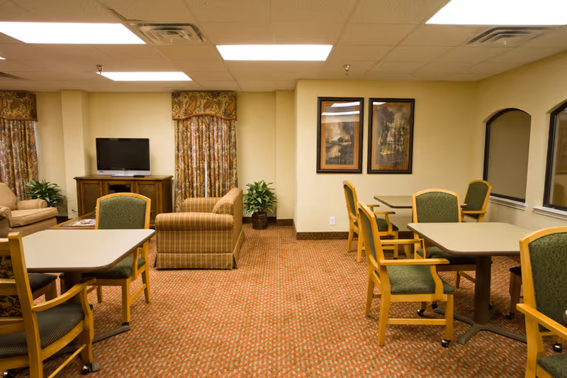 A common area in an assisted living facility featuring several tables with green cushioned chairs, a seating area with two armchairs and a loveseat, a TV on a wooden stand, patterned curtains, two framed paintings on the wall, and potted plants in the corners.