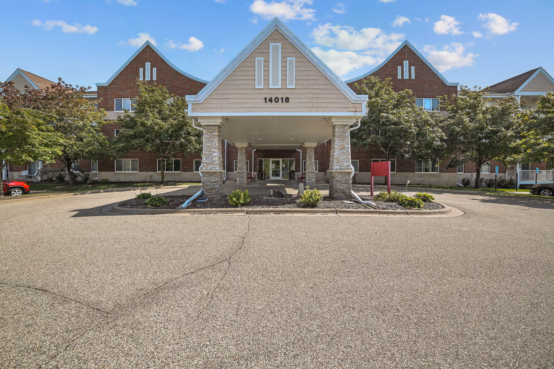 Front exterior view of a senior living facility building with a covered entrance supported by stone pillars, surrounded by trees and a parking area under a blue sky with some clouds. The building has a brick facade with beige siding on the entrance gable and the number 14018 displayed above the entrance.