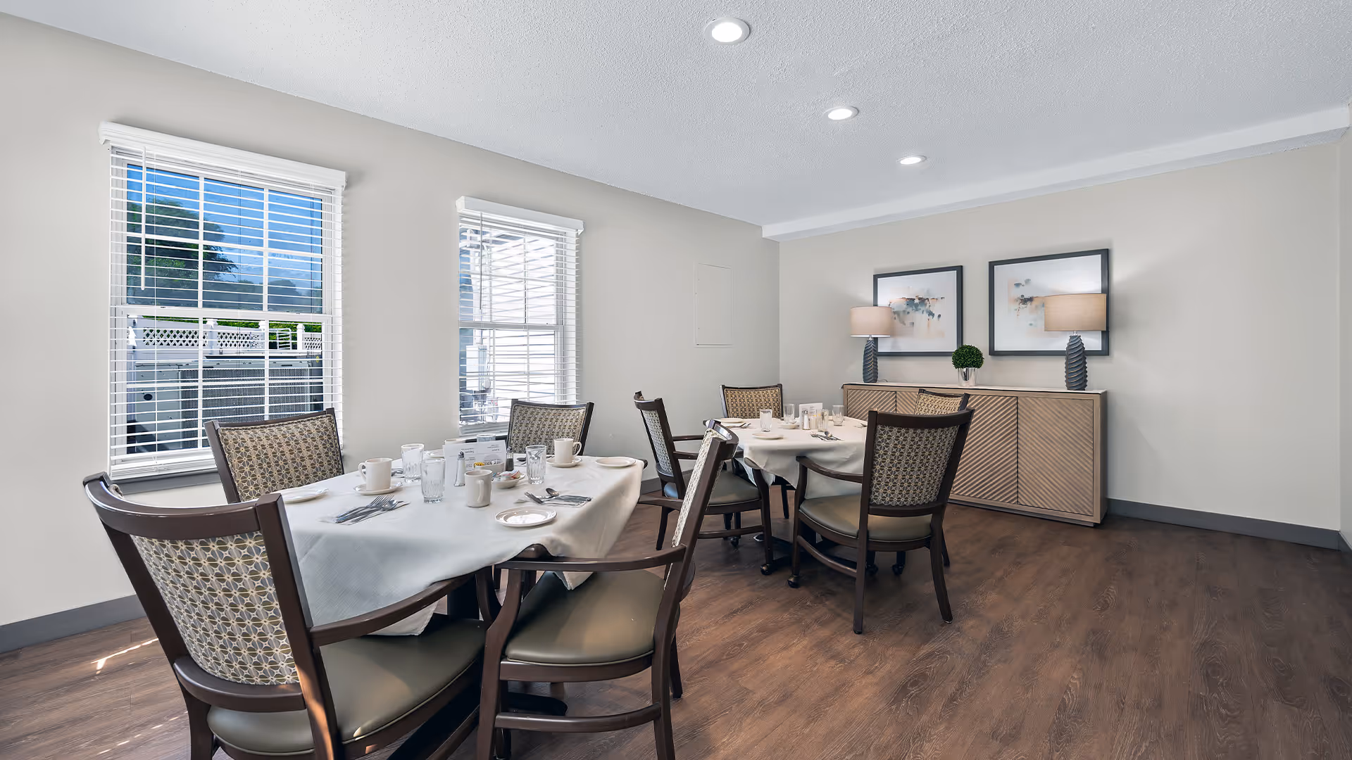 A dining room with two tables covered with white tablecloths, set with cups, glasses, plates, and silverware. Six chairs surround the tables. The room has two windows with white blinds, wooden flooring, and a sideboard with two lamps and framed artwork above it.