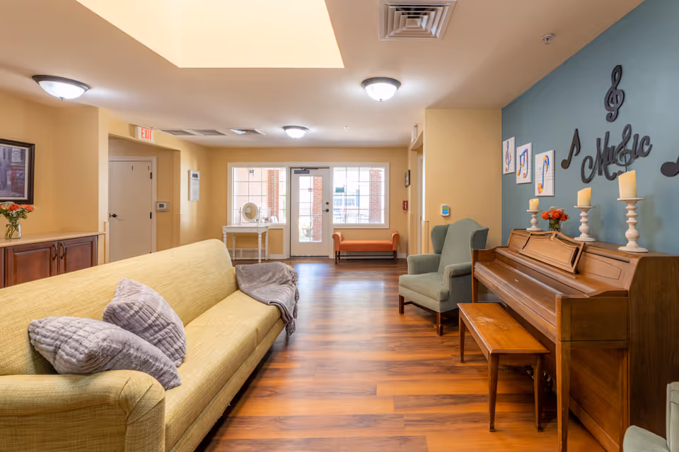 A cozy common area in a senior living facility featuring a yellow sofa with gray pillows and a gray throw, a green armchair, a wooden piano with candles and flowers on top, and musical decorations on a blue accent wall. The room has wooden flooring, ceiling lights, and a door with windows leading outside.