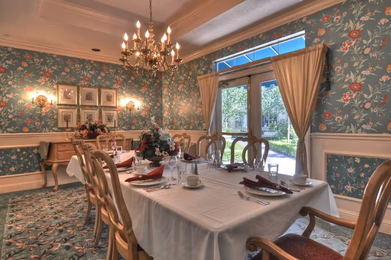 A formal dining room with a long table covered with a white tablecloth set with plates, glasses, cups, and folded napkins. The room has floral wallpaper, a chandelier hanging from the ceiling, framed pictures on the wall, and a window with curtains showing a garden outside.