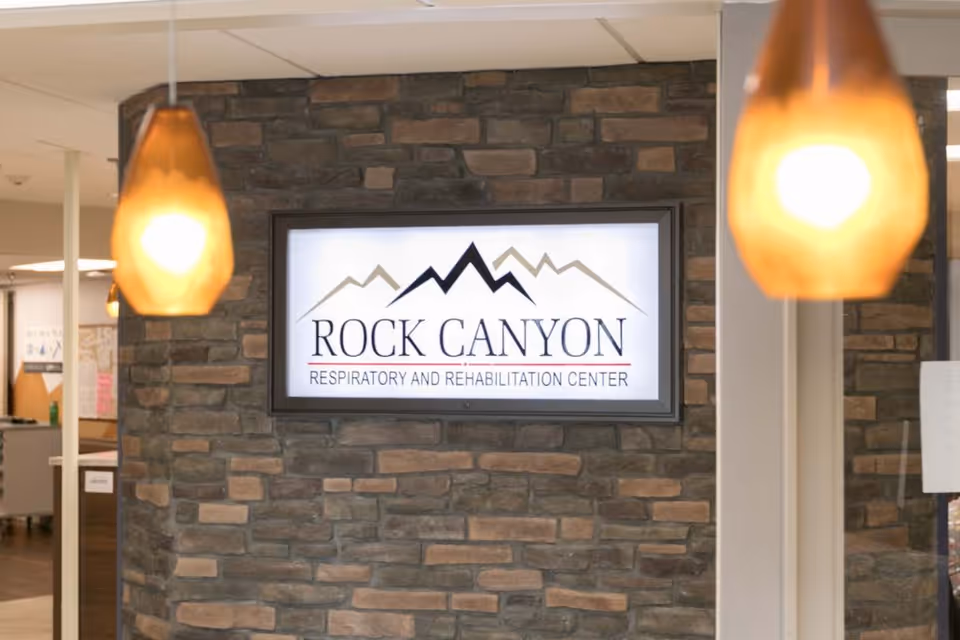 A lit sign mounted on a stone wall inside a building reads 'Rock Canyon Respiratory and Rehabilitation Center' with a mountain logo above the text. Two hanging amber pendant lights are visible in the foreground.