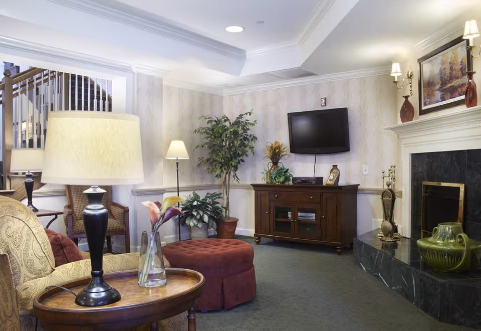 A cozy living room area with a patterned armchair, a round wooden side table with a lamp and a vase of flowers, a burgundy ottoman, a wooden TV stand with a flat-screen TV mounted on the wall above it, potted plants, a fireplace with decorative items on the mantel, and a staircase in the background.
