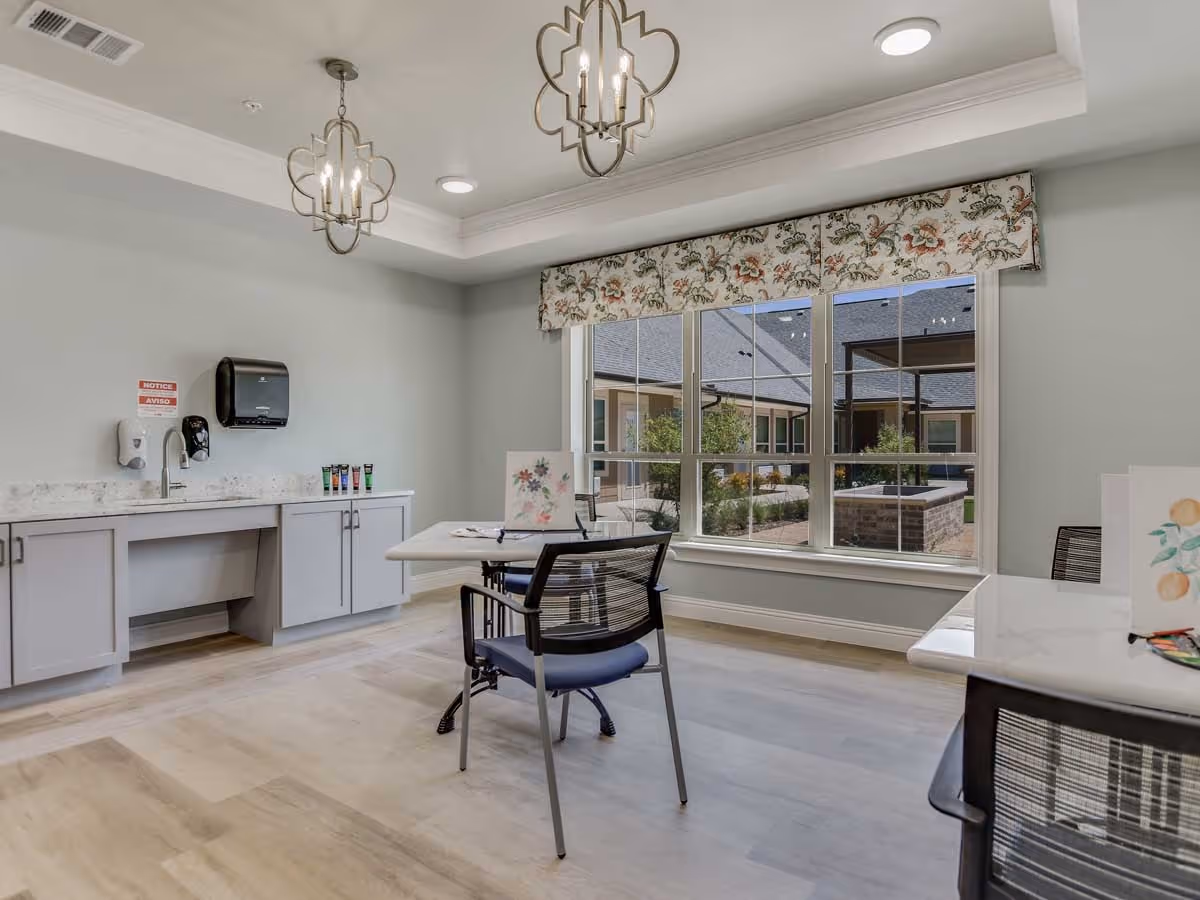 Bright activity room with tables and chairs, a countertop with sink and supplies, and a large window overlooking an outdoor courtyard.