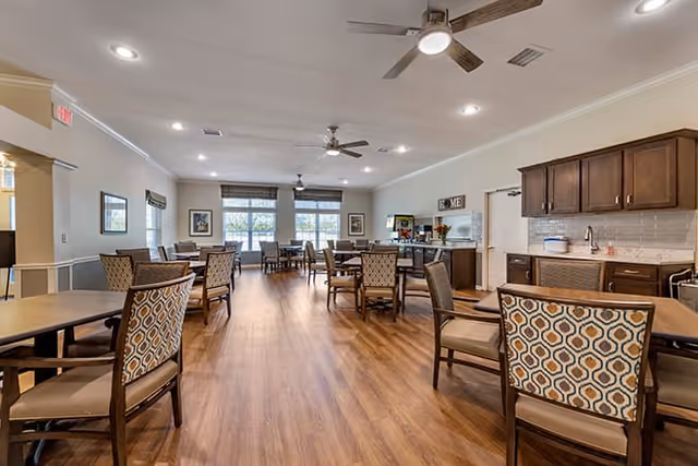 A spacious dining room with wooden flooring, multiple tables and chairs with patterned upholstery, ceiling fans, and large windows letting in natural light. There is a kitchenette area with cabinets, a sink, and a coffee station in the background.