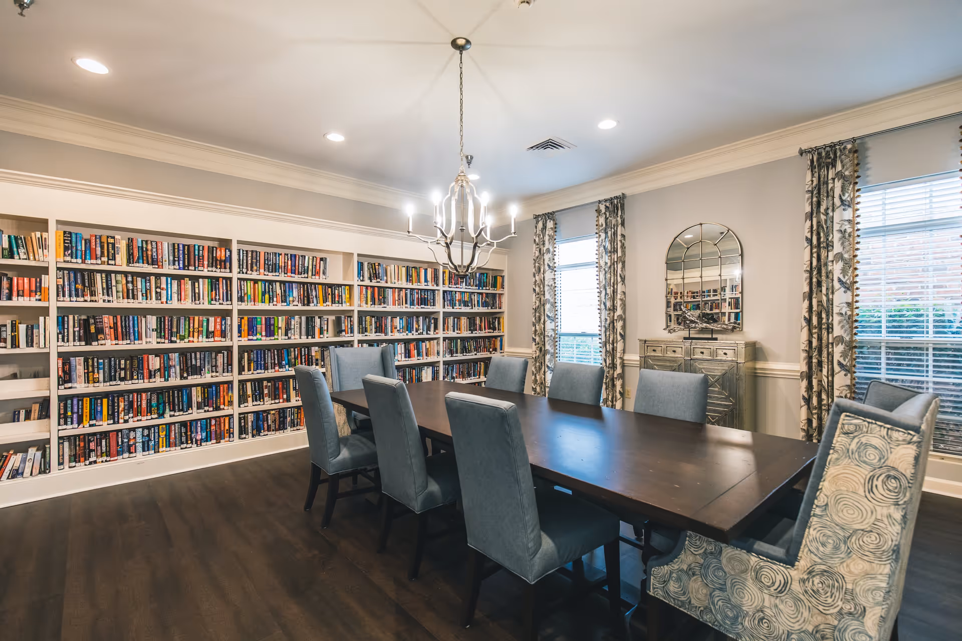 A well-lit room with a long dark wooden table surrounded by eight upholstered chairs, some in solid blue and others with a blue and beige swirl pattern. The room features a large bookshelf filled with books along one wall, two windows with floral curtains, a decorative mirror above a small cabinet, and a chandelier hanging from the ceiling.