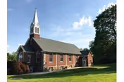 A red brick church building with a white steeple and multiple windows, situated on a well-maintained grassy lawn with trees in the background under a partly cloudy blue sky.