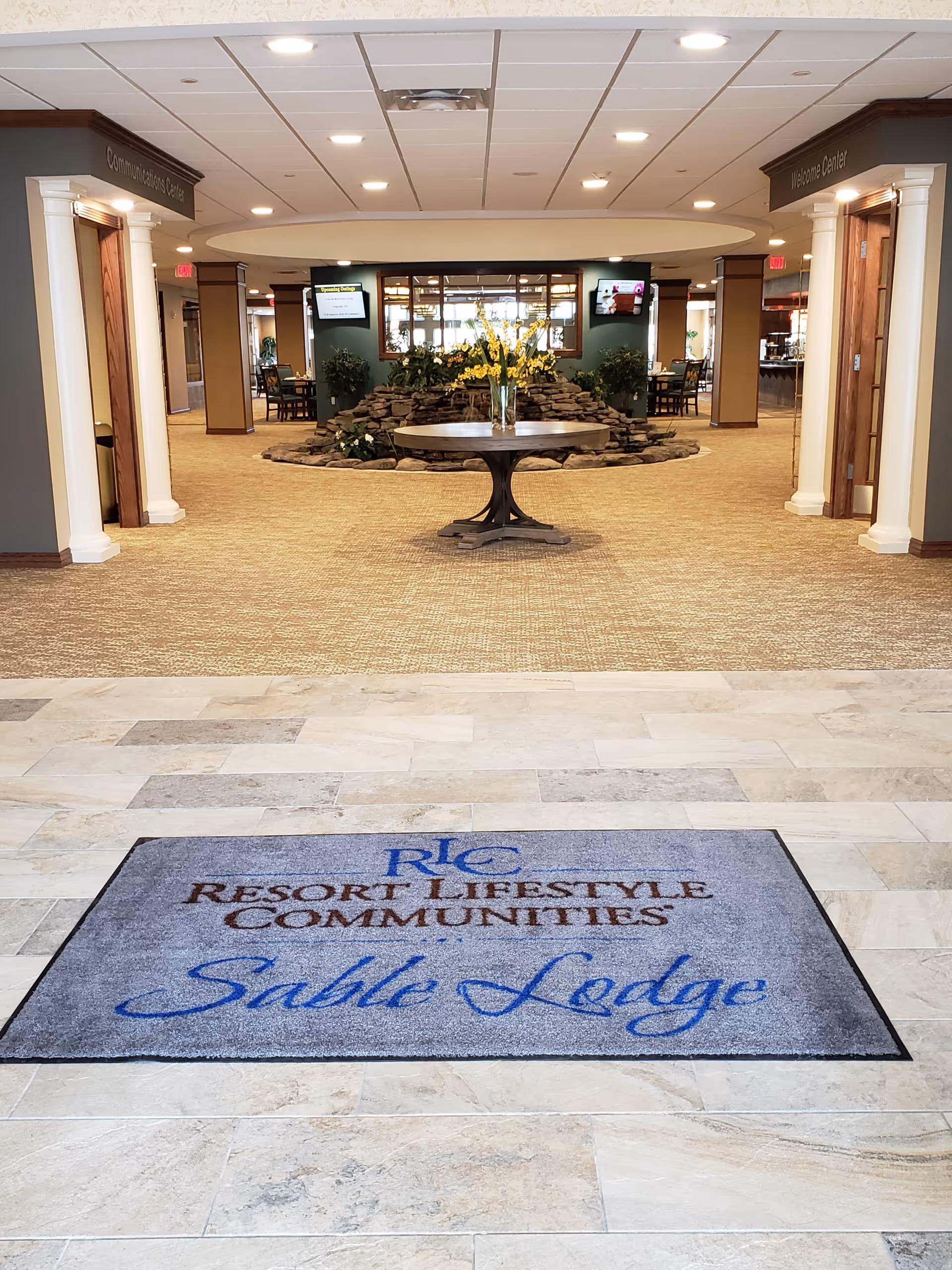 Entrance area of Sable Lodge Retirement Community featuring a floor mat with the community's name, a round table with a vase of yellow flowers, stone water feature, and signs for Community Activities Center and Welcome Center on either side.