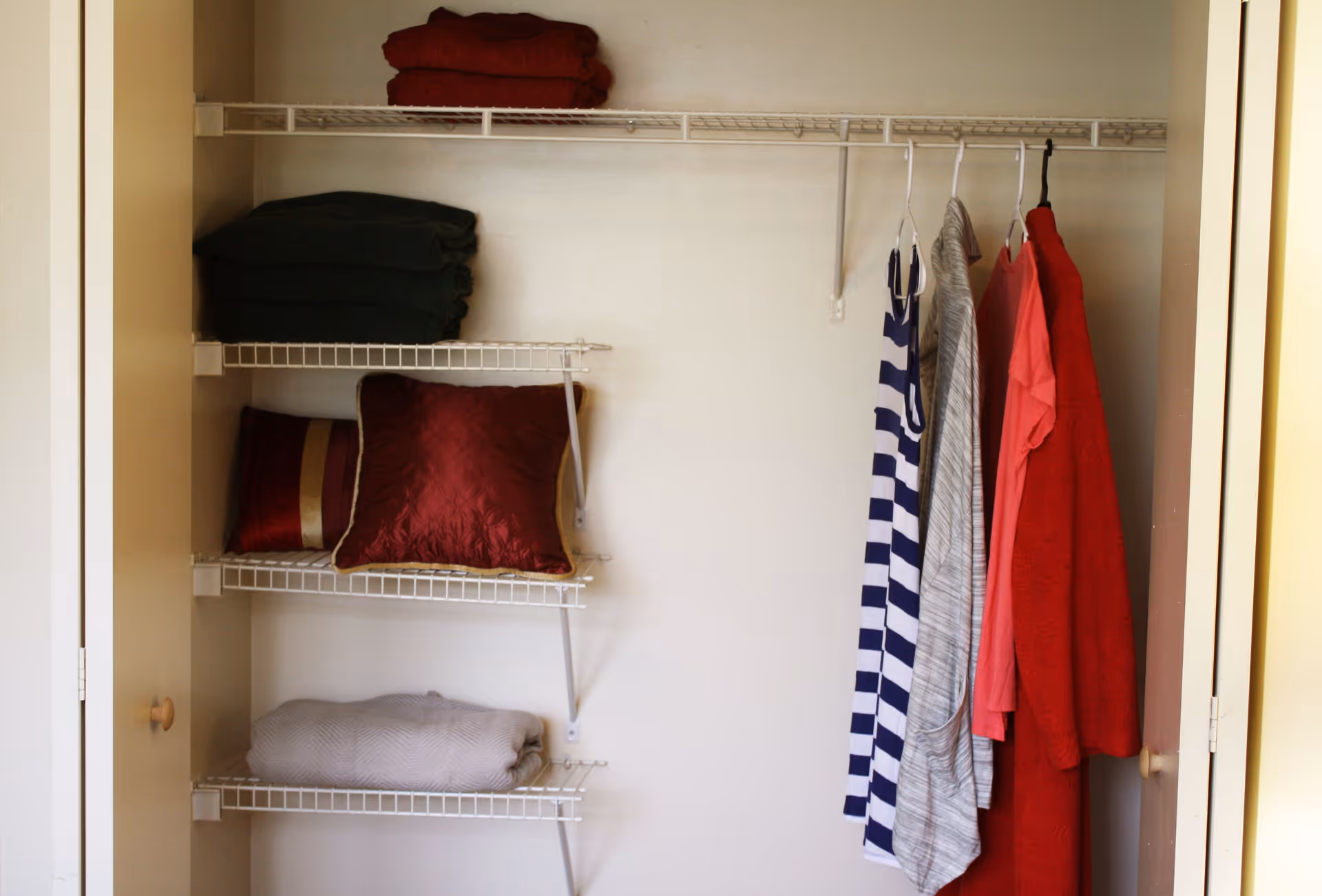 An open closet with wire shelves holding folded blankets and pillows on the left side, and several hanging clothes including a striped dress, a gray garment, a pink shirt, and a red coat on the right side.