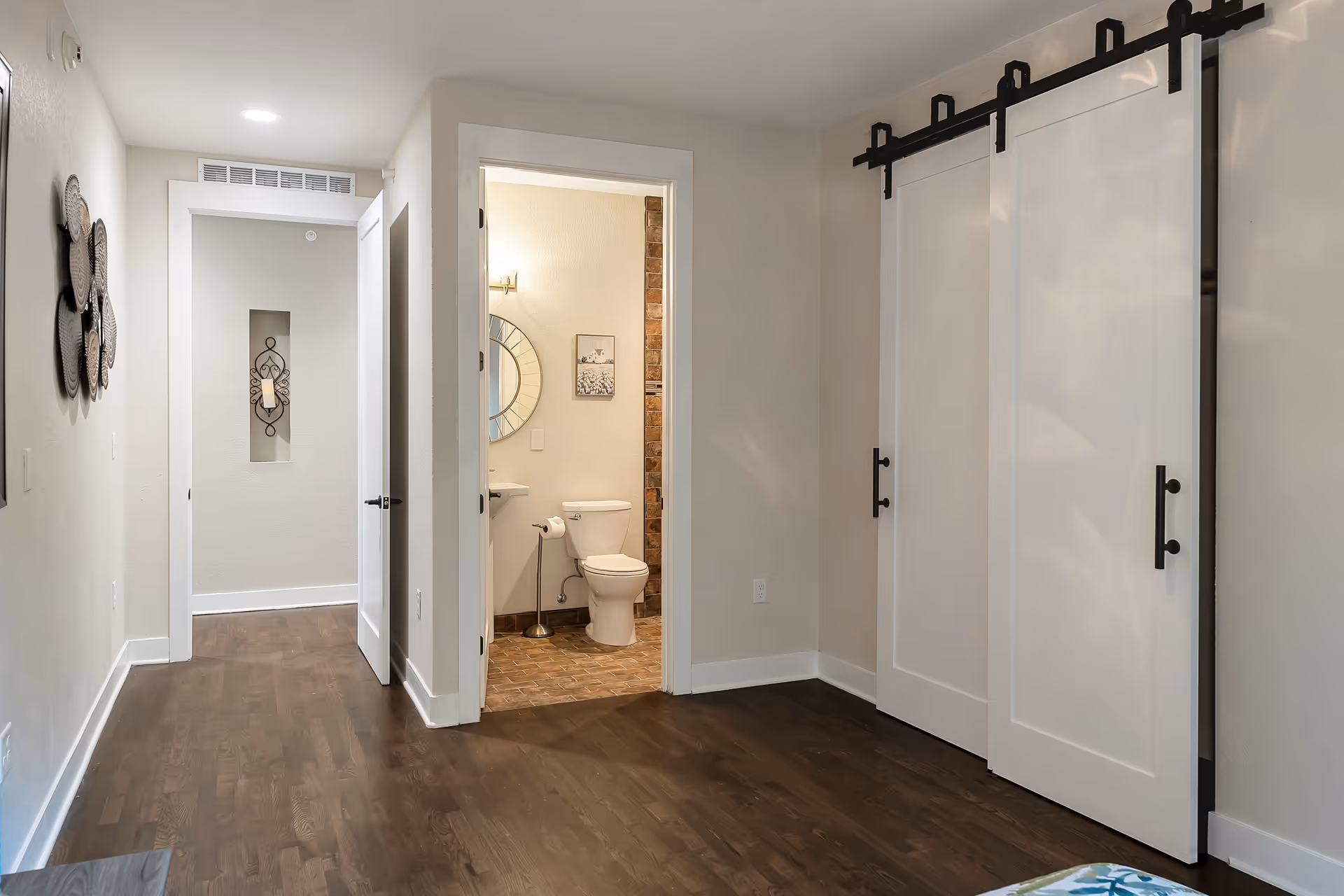 Interior view of a senior living facility room with wooden flooring, a hallway leading to a bathroom with a toilet and a round mirror above the sink. The room features white sliding barn doors on a black rail and neutral-colored walls with decorative wall art.